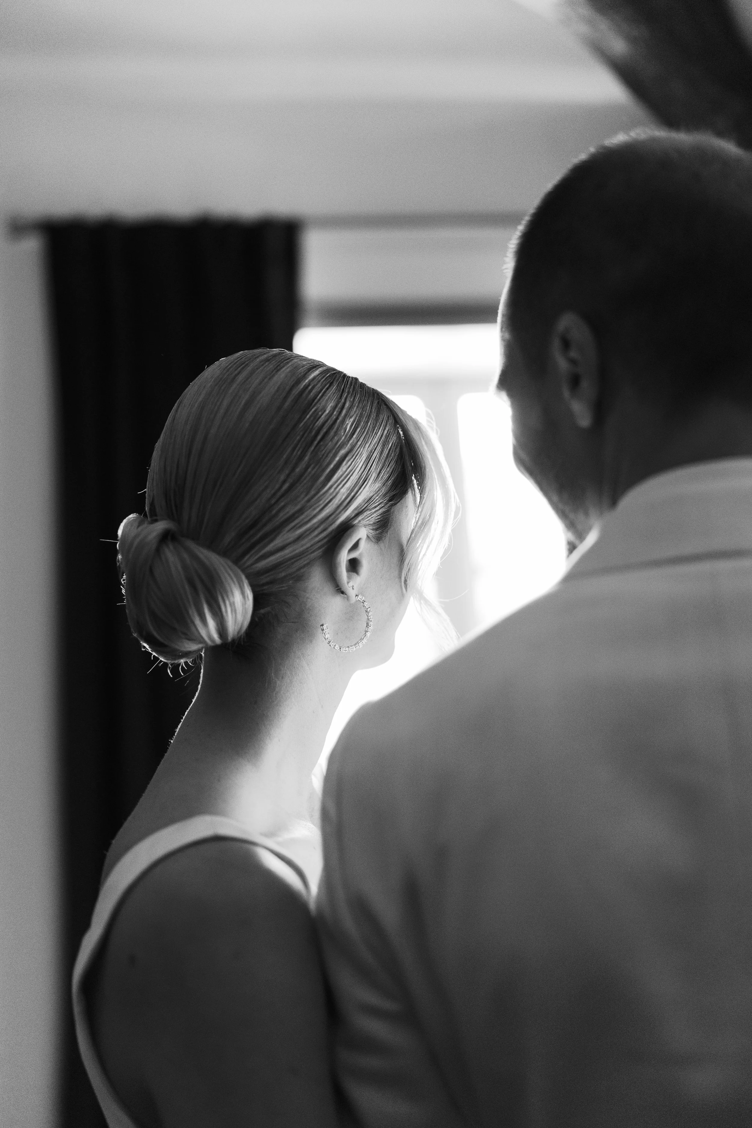 A woman with styled hair and hoop earrings standing close to a man, both facing away from the camera, near a window with curtains, in black and white.