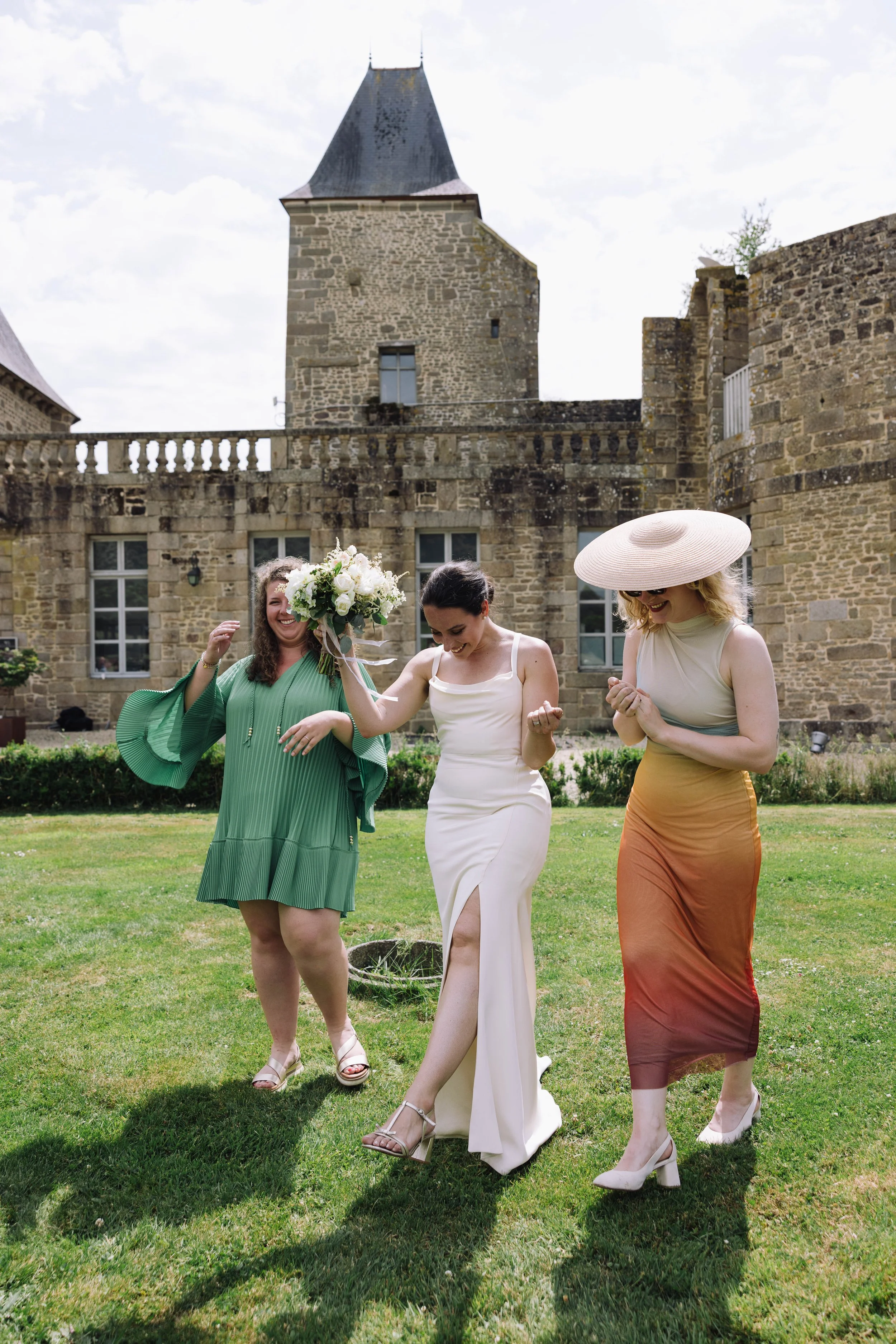 Three women celebrating outdoors in front of an old stone castle, one holding a bouquet of flowers, all smiling and happily dancing.