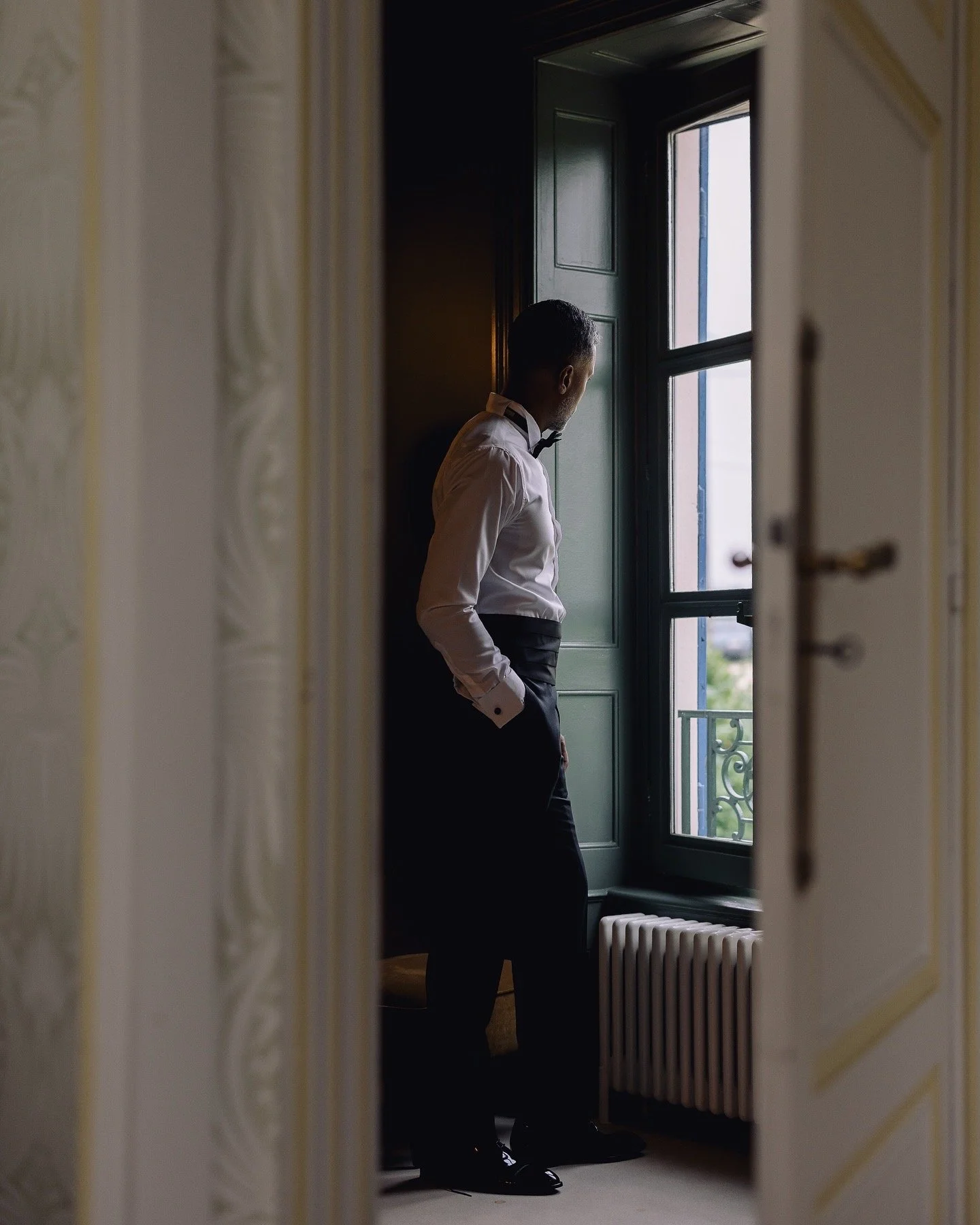 A man in a tuxedo with a bow tie looking out of a window in a room with decorative molding.