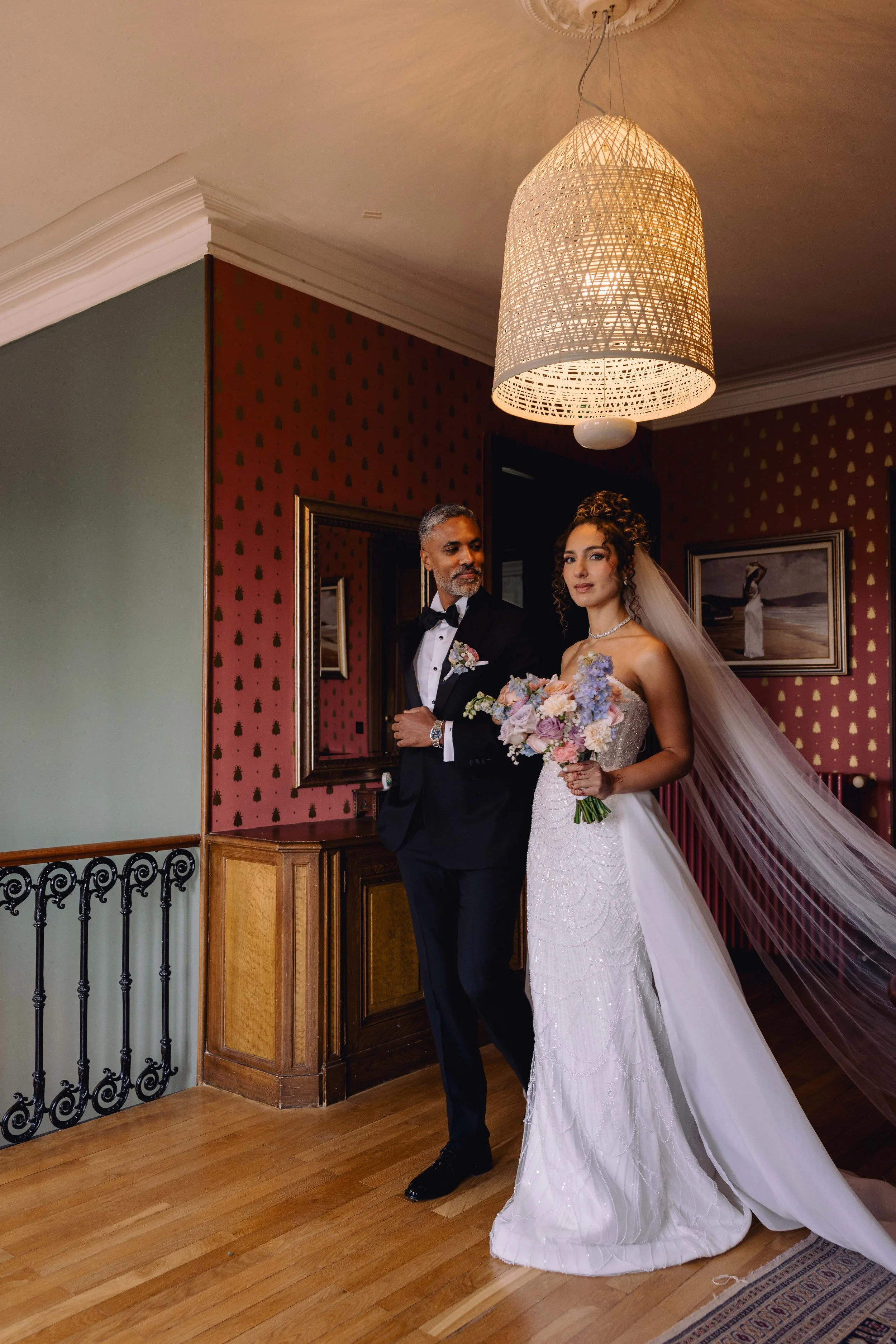 A bride holding a bouquet of flowers standing next to a man in a tuxedo inside a decorated room with wooden floor, patterned wallpaper, framed pictures, and a large hanging lamp.