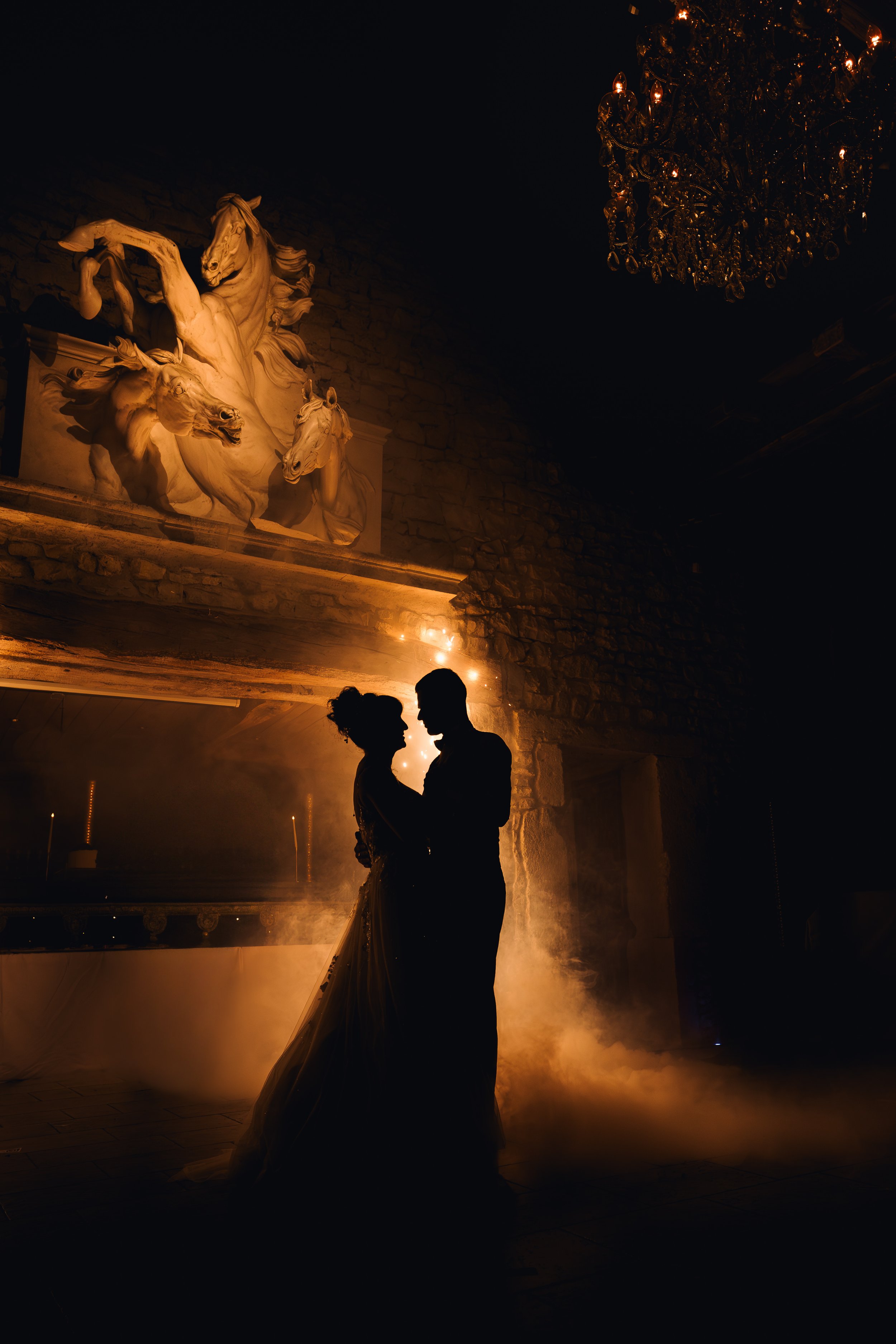 Silhouetted couple dancing indoors with warm lighting, stone wall, ornate sculpture of horses, and chandelier in background.