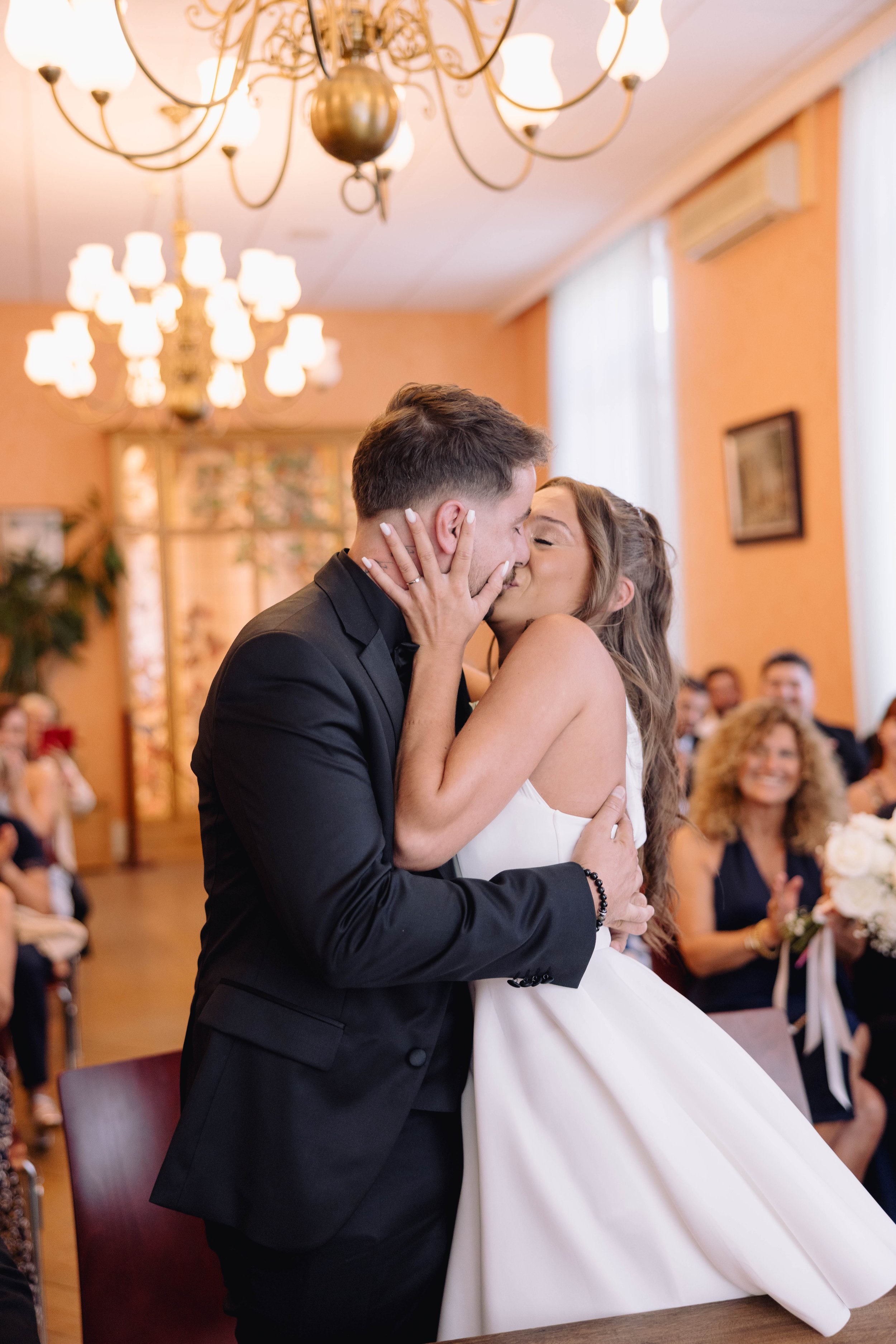A bride and groom sharing a kiss during their wedding ceremony, surrounded by smiling guests in a decorated indoor venue with warm lighting.