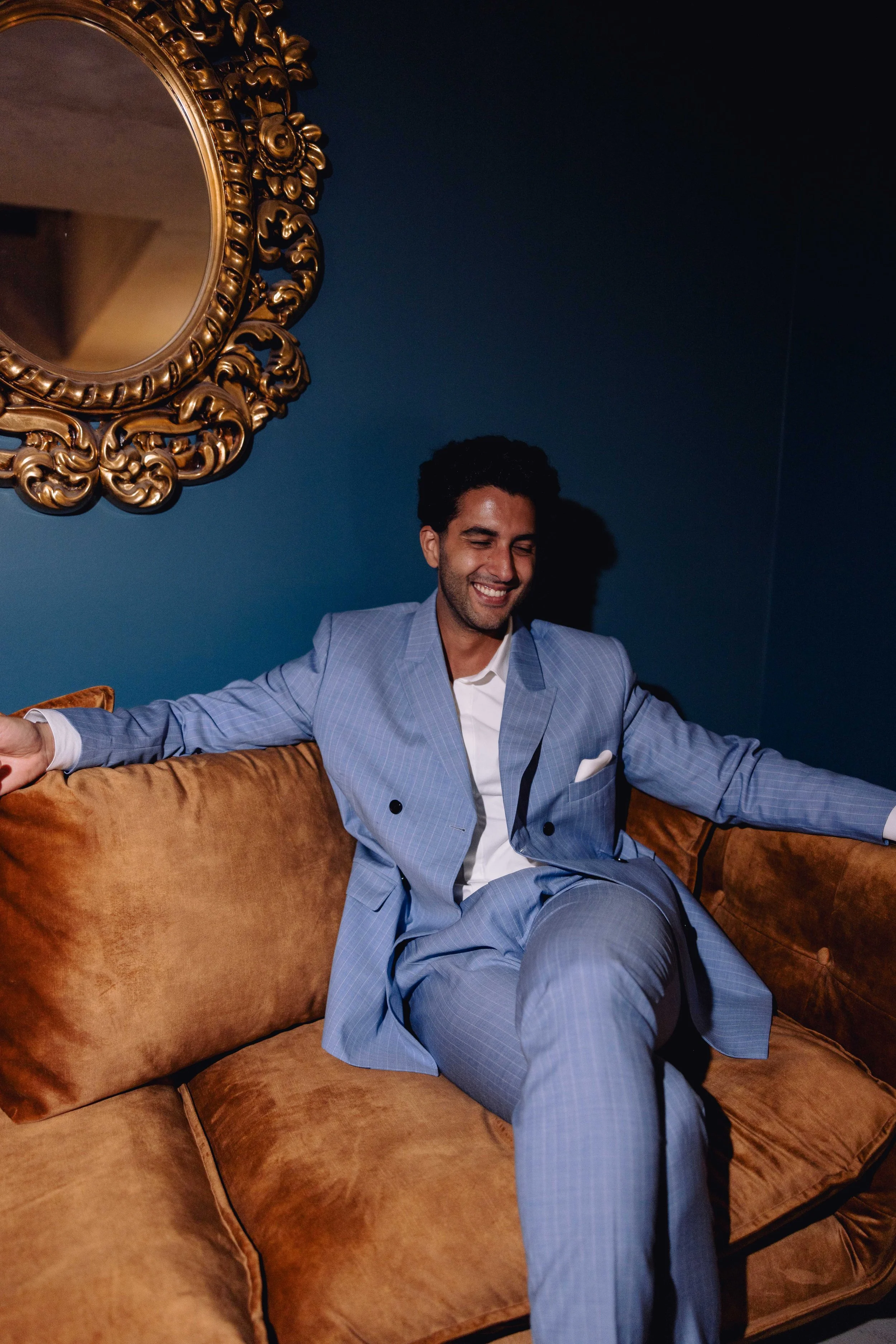 A smiling man in a light blue suit sitting on a brown velvet sofa in a room with dark blue walls and a gold ornate mirror.