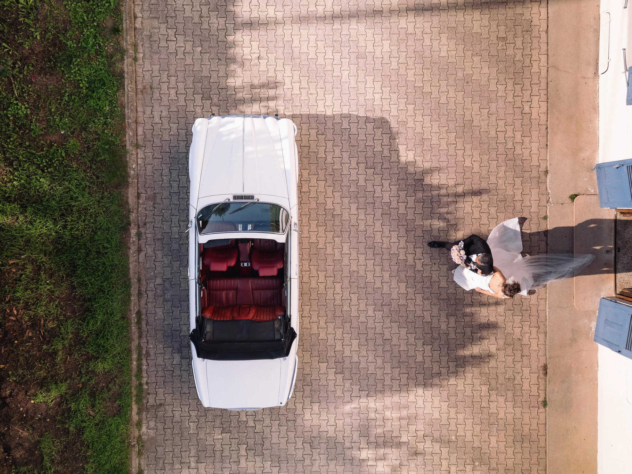 A white convertible car parked on a brick pavement next to a woman in a wedding dress and a man in formal attire, possibly a bride and groom, seen from above.