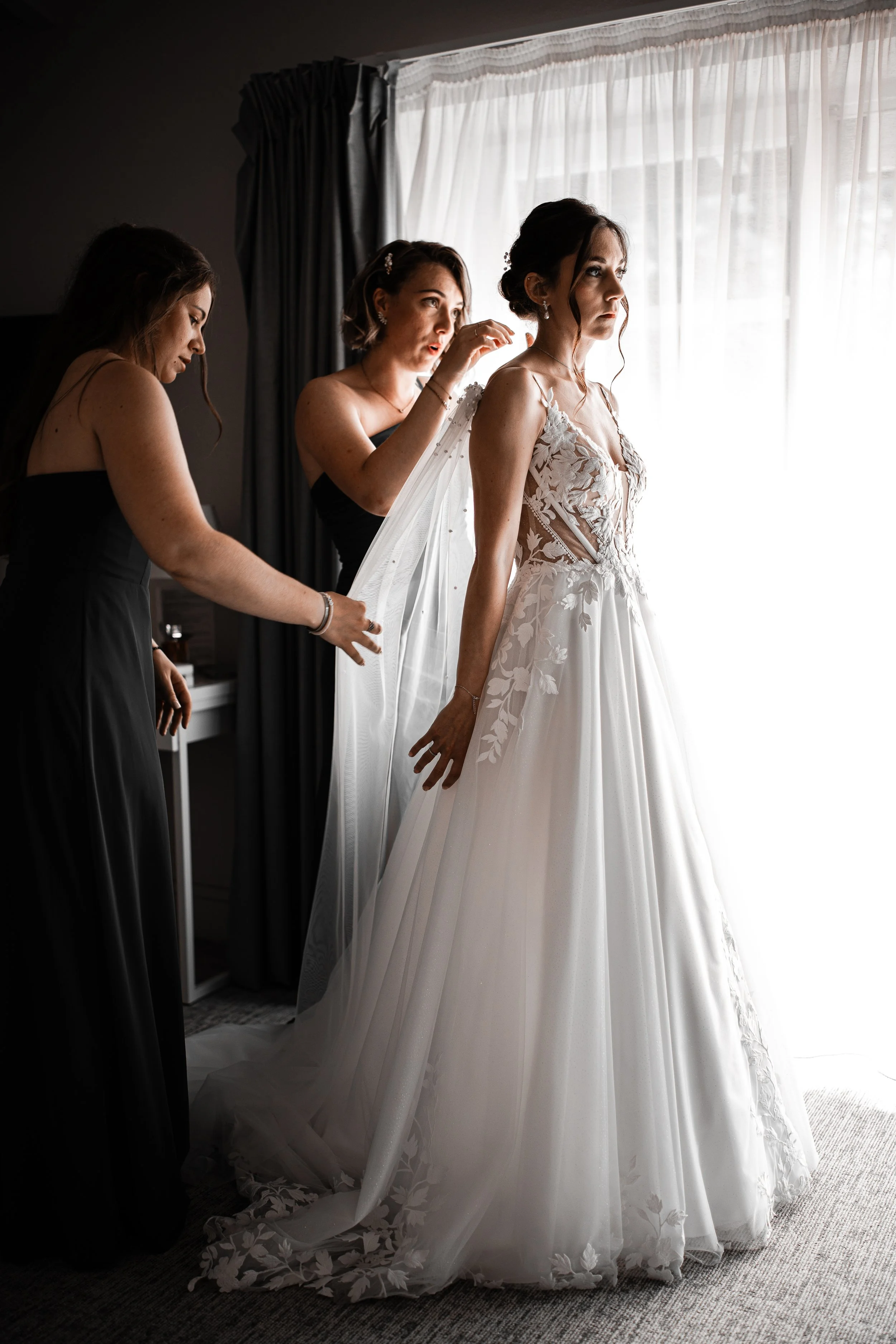 Bride in a white wedding dress standing by a window as two women assist her with her veil.