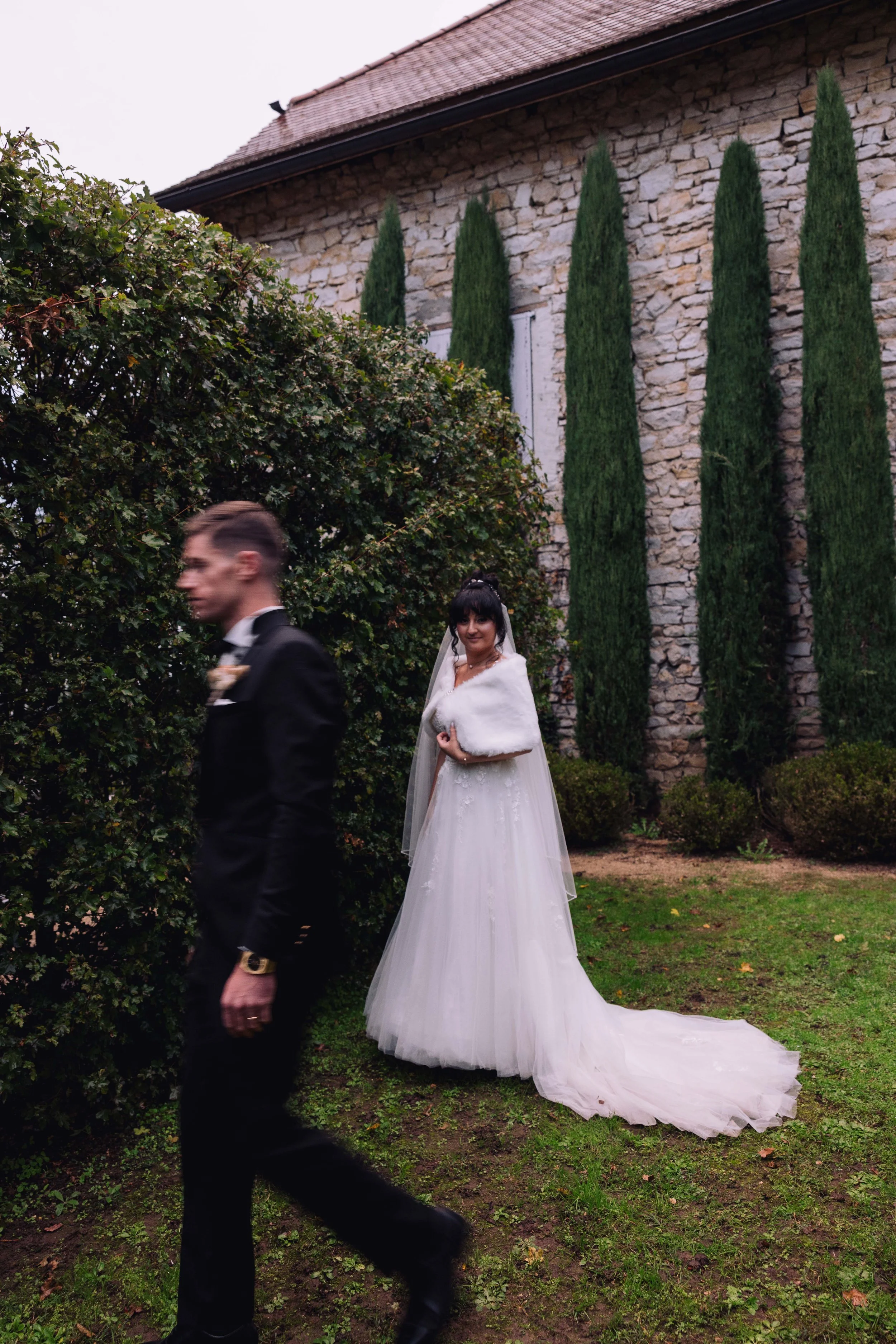 Bride in a white wedding gown and fur stole stands near a stone building with tall, narrow trees in the background, while a man in a black suit walks past in the foreground.
