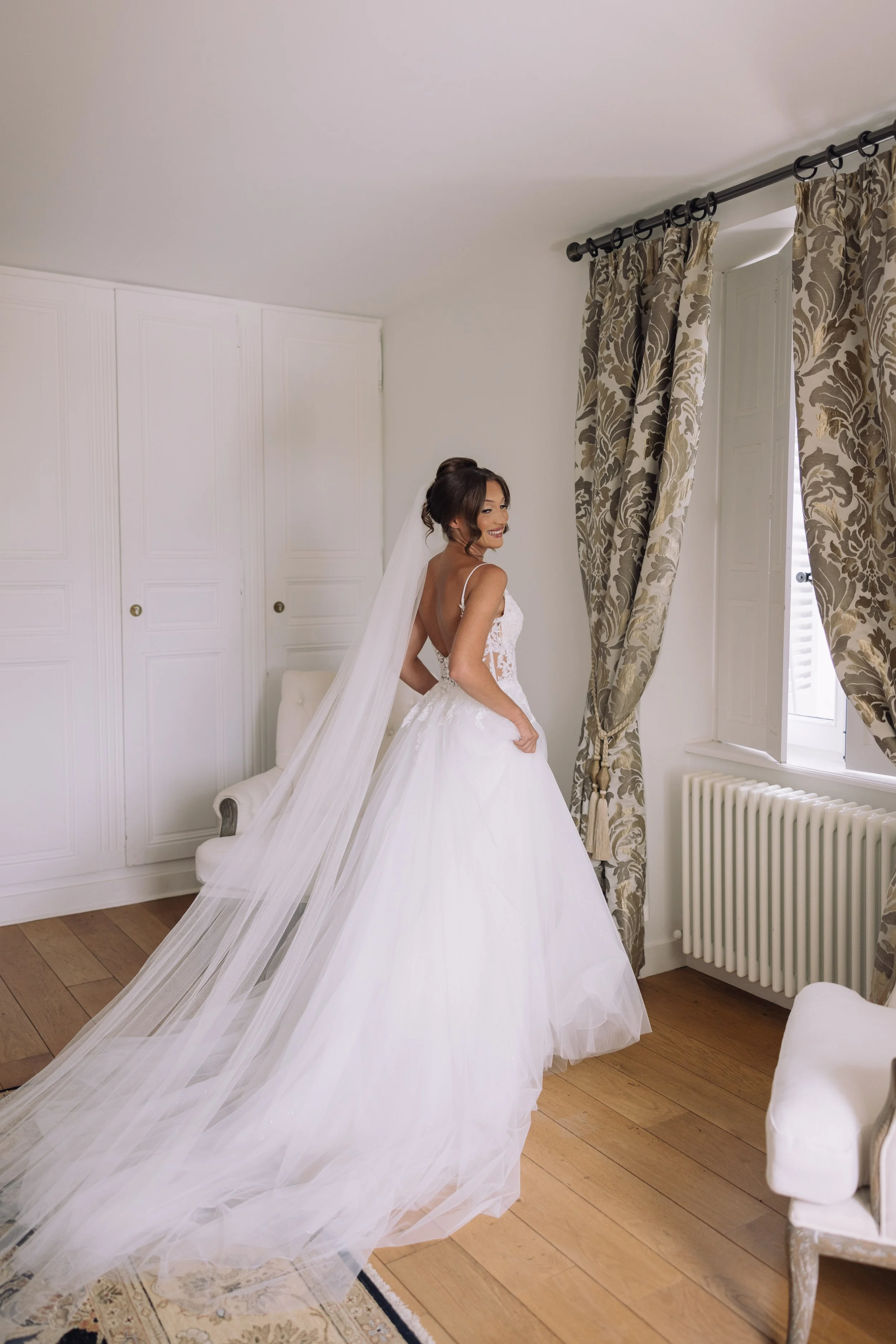 A bride in a white wedding gown and long veil standing in a well-lit room with wooden floors, patterned curtains, and a white chair, looking over her shoulder and smiling.