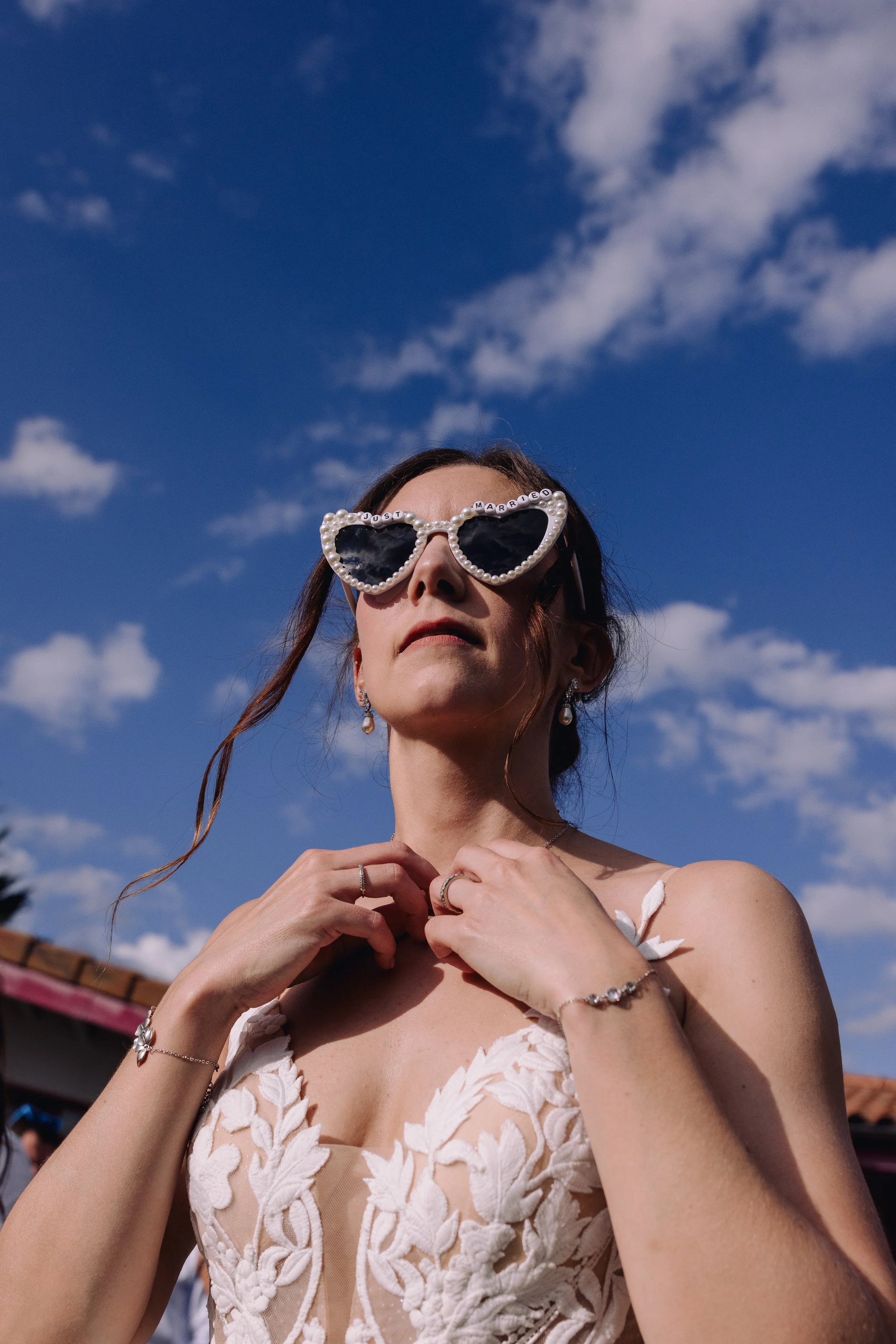 A woman wearing heart-shaped sunglasses with pearl frames and the words "Just Married" on the top, dressed in a white lace dress, standing outdoors against a blue sky with scattered clouds, adjusting her necklace.