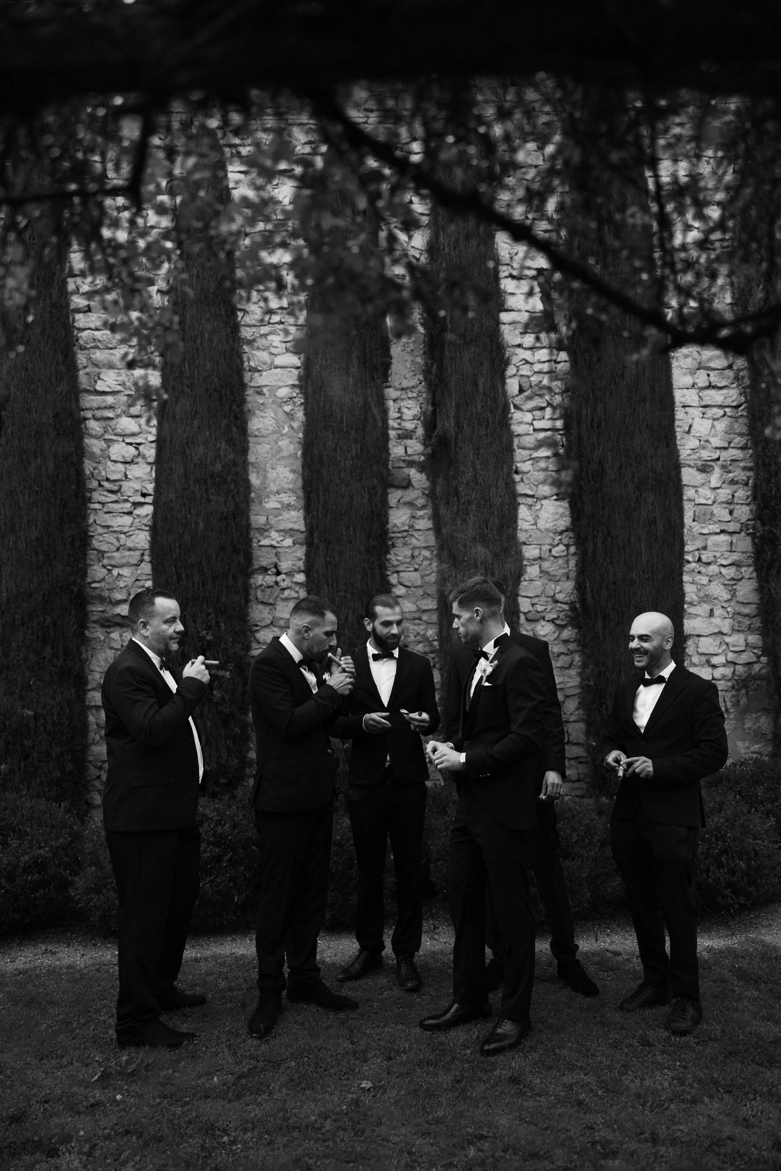 Six men in tuxedos standing in a garden, having a conversation, with a brick wall and tall trees in the background.