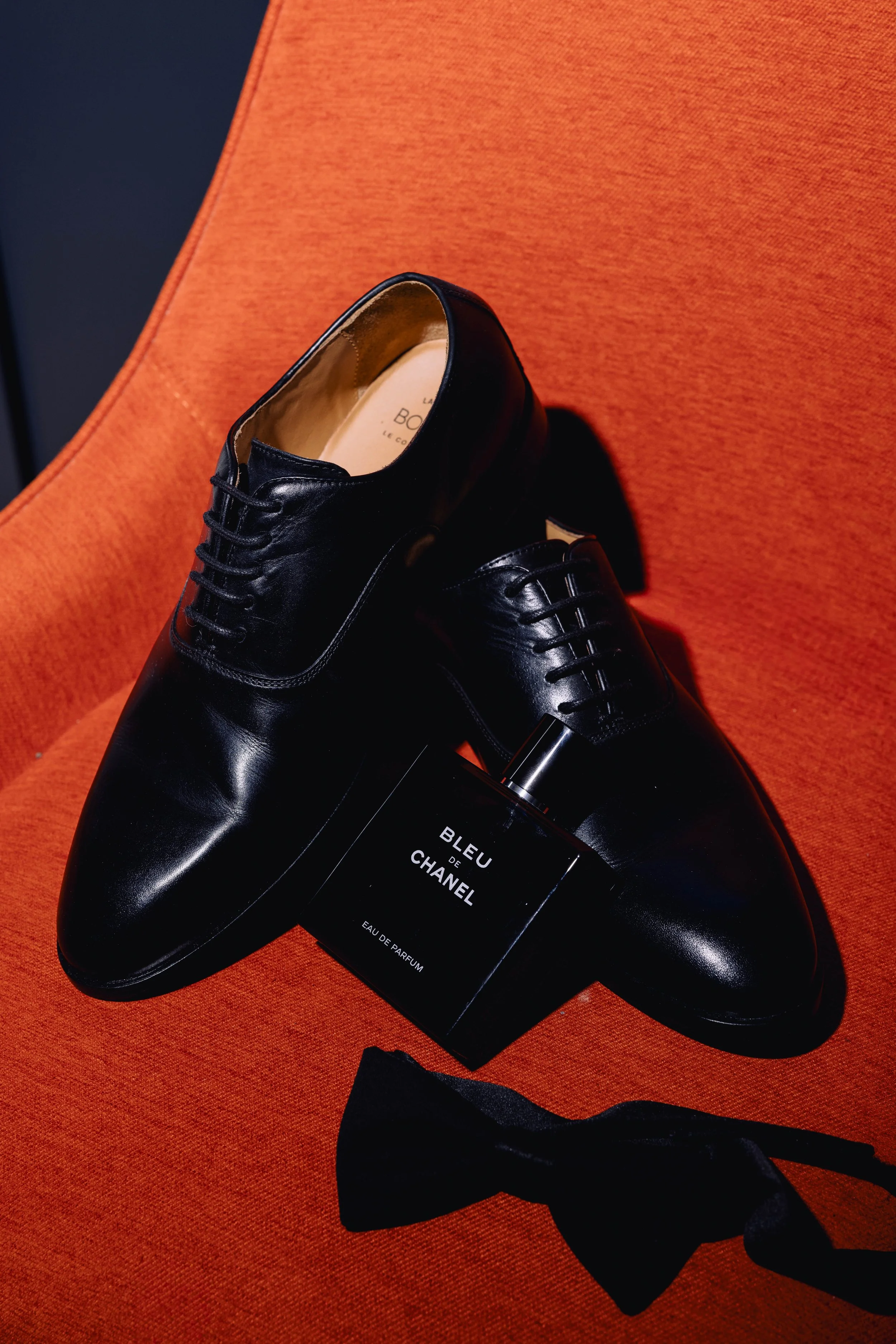 A pair of black leather dress shoes, a black Chanel Bleu de Chanel perfume bottle, and a black bow tie on an orange upholstered surface.