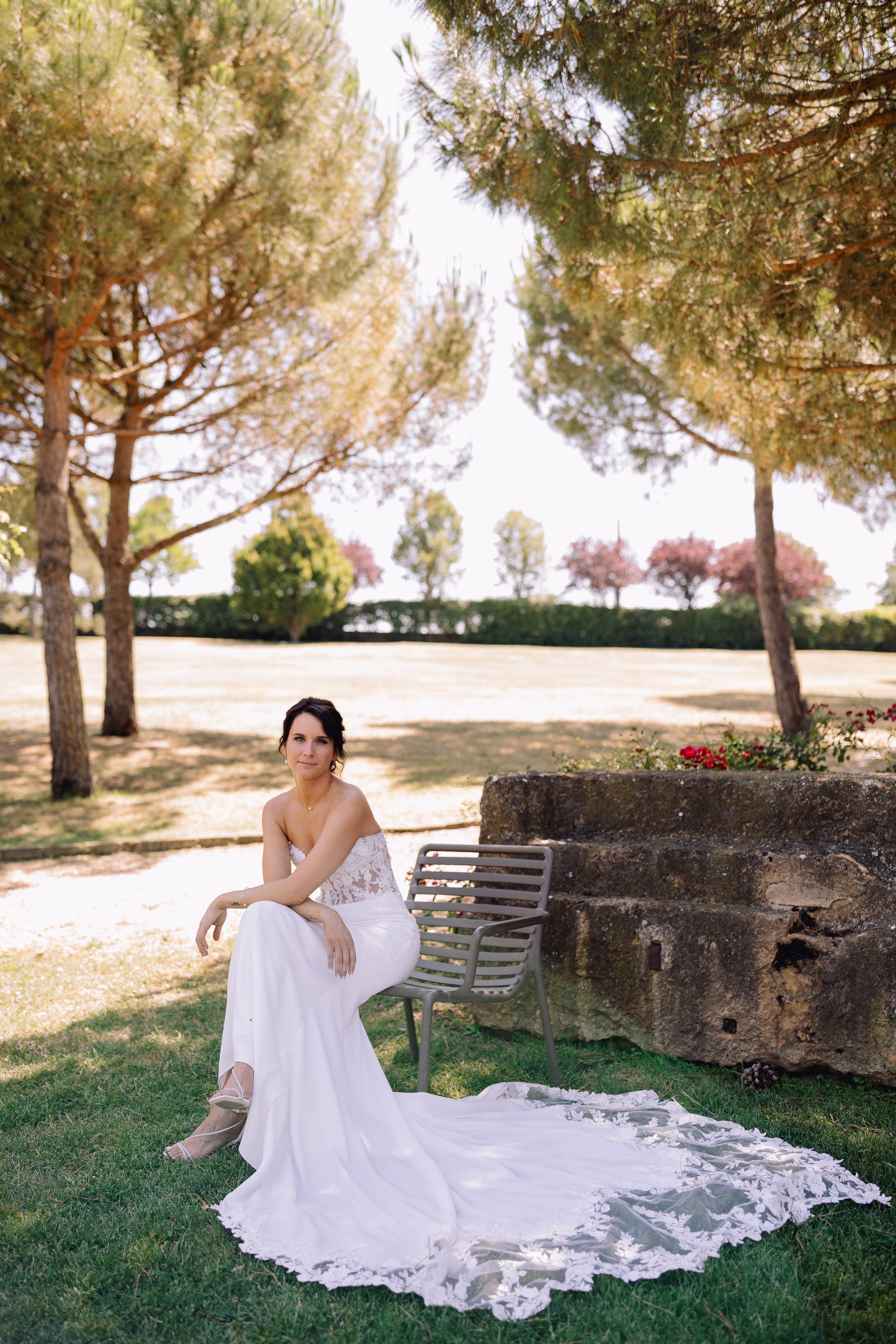 A woman in a white wedding dress sitting on a bench outdoors, with trees and grass in the background.