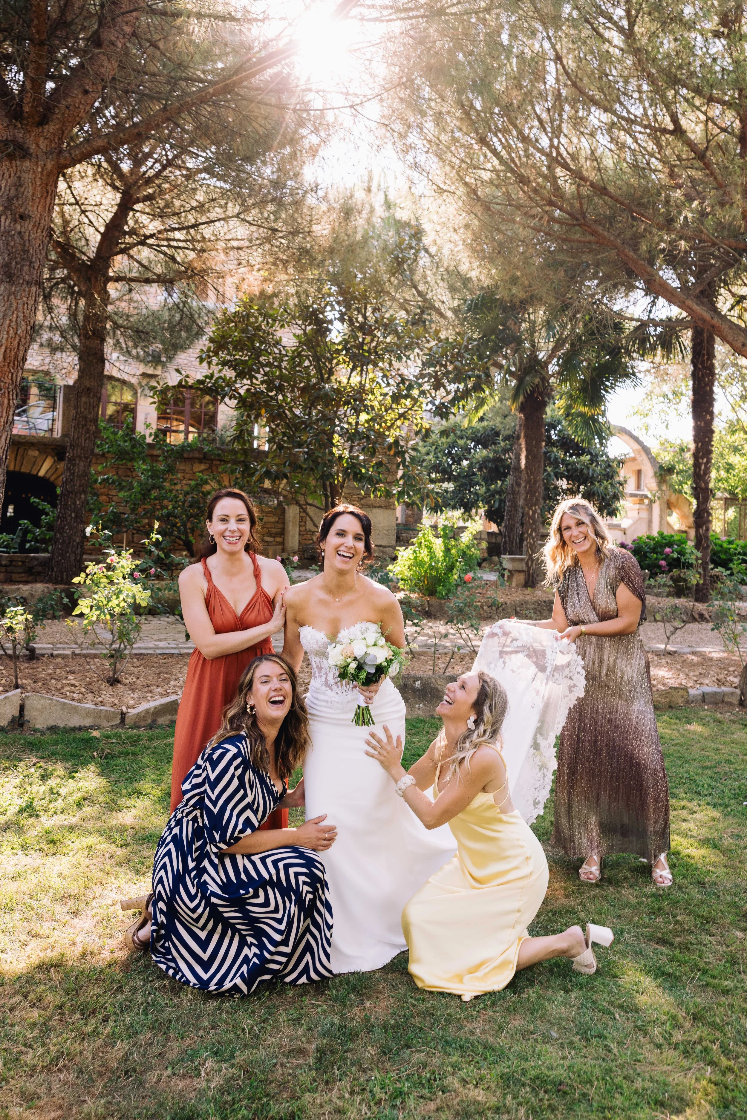 A bride and her five bridesmaids celebrating outdoors on her wedding day with sunlight filtering through trees in the background.