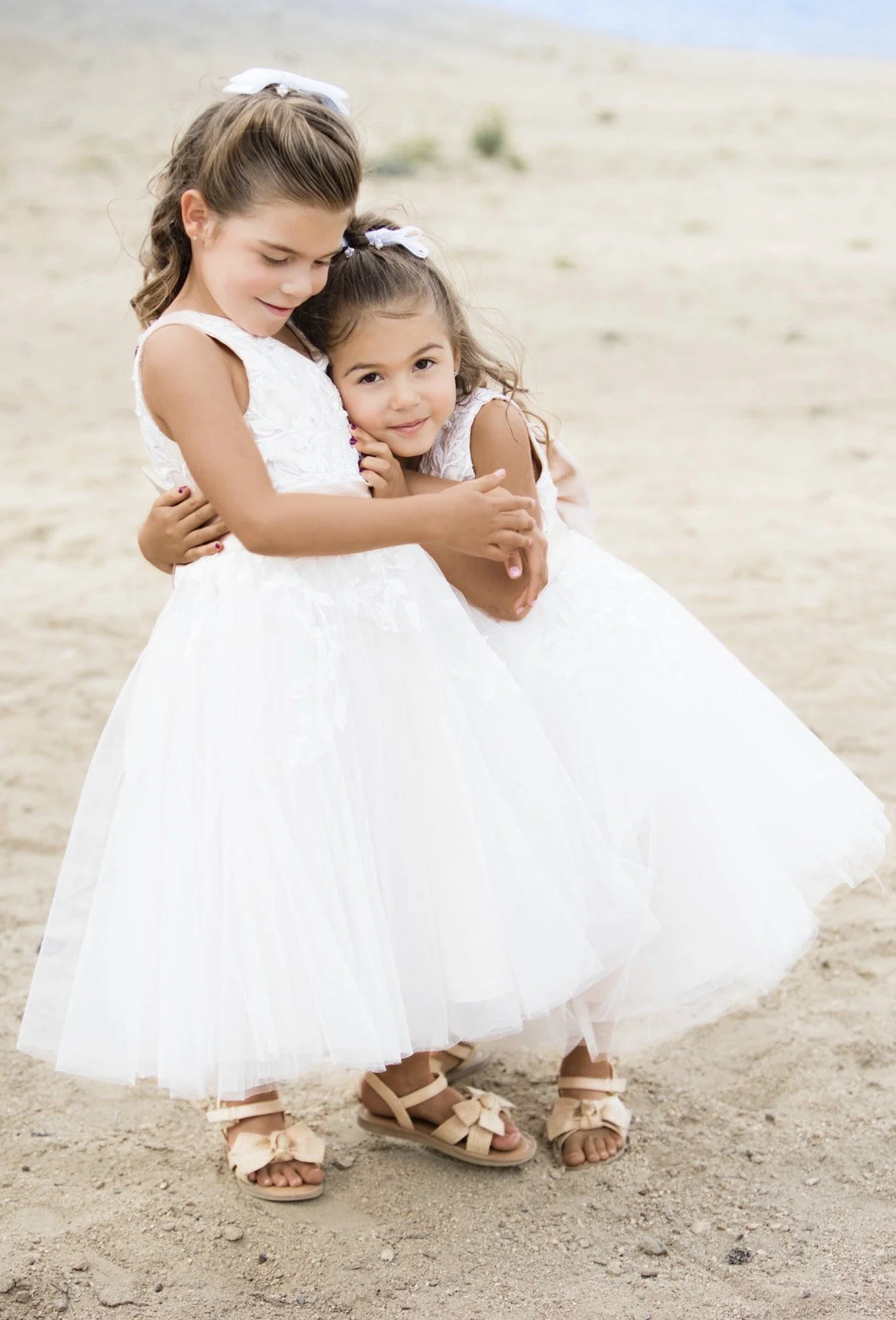 Two young girls in white dresses hugging each other on a sandy beach, wearing sandals and white bows in their hair.