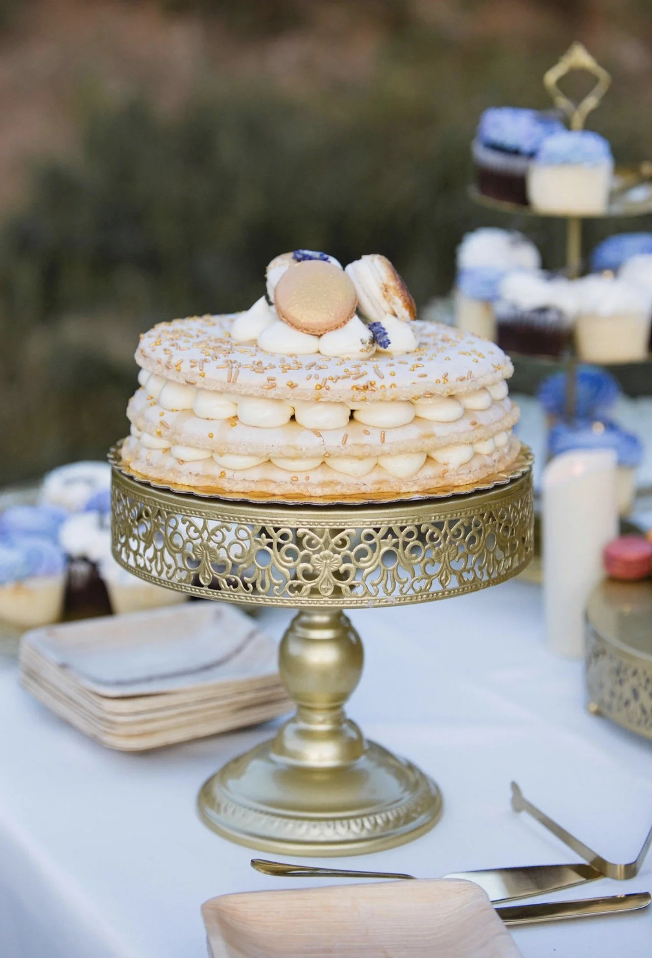 A layered white and gold cake with decorative toppings on a gold ornate cake stand, with smaller desserts in the background.