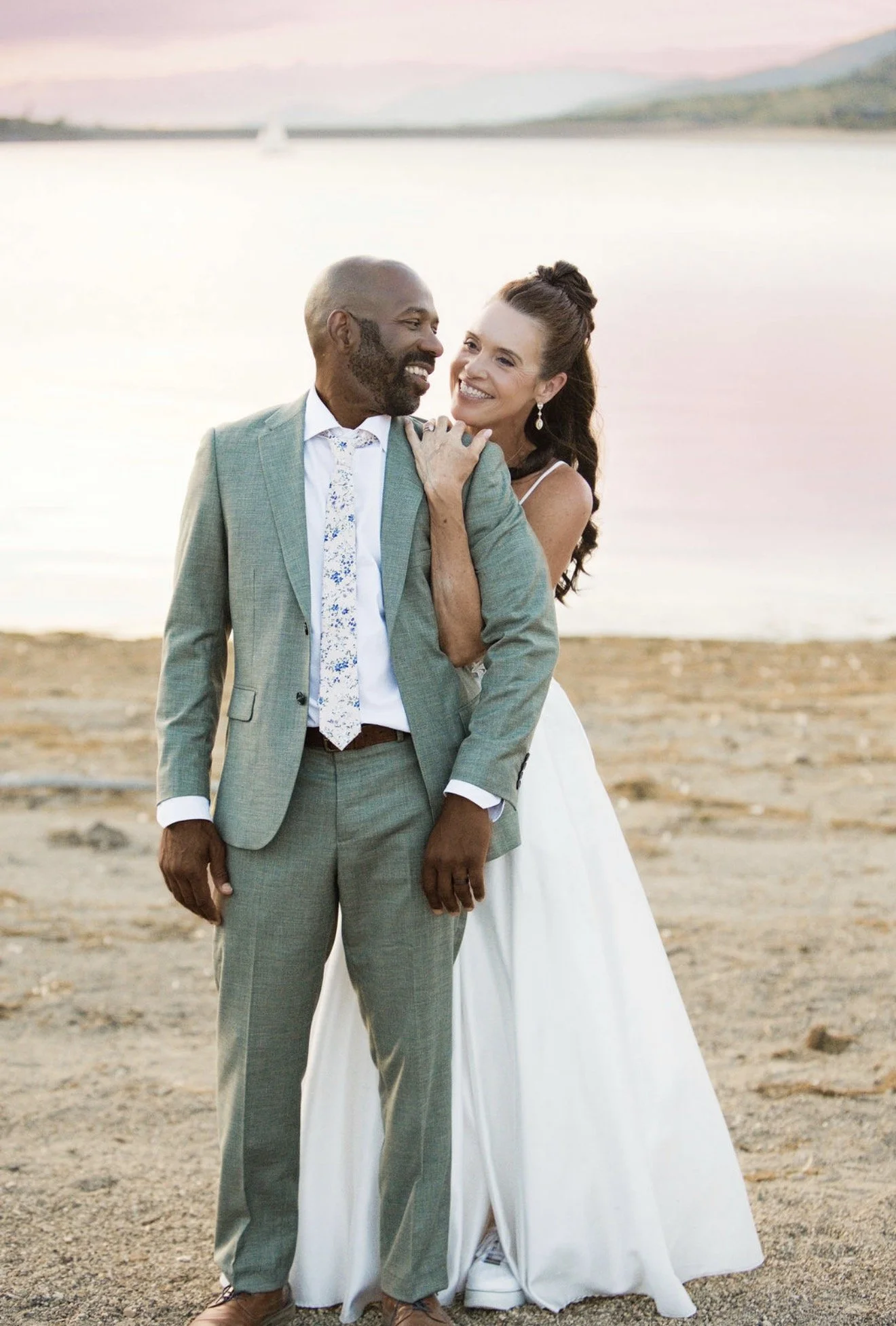 A smiling couple, a man and a woman, standing close together on a beach at sunset, with a body of water and mountains in the background. The man is wearing a light green suit and the woman is in a white dress.