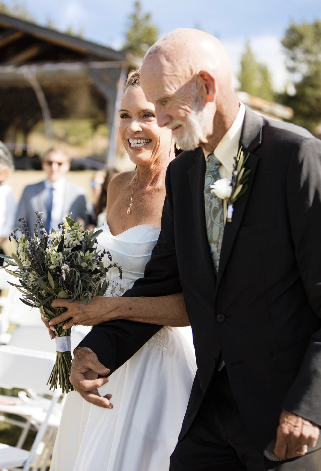 A smiling bride holding a bouquet of flowers walking with an elderly man at an outdoor wedding ceremony.