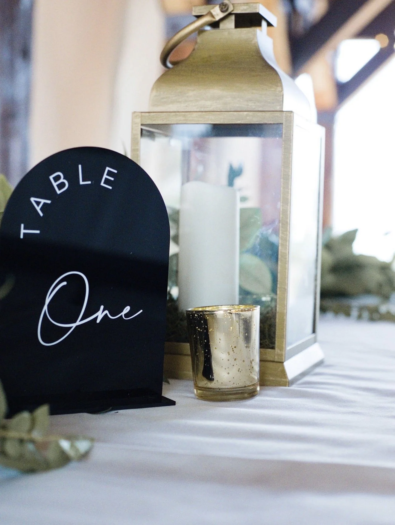 A black decorative sign with white cursive text saying "Table One" positioned on a white table. Behind it is a gold and glass lantern-style candle holder with a white candle inside and a gold-colored candle in front of the lantern. In the background,