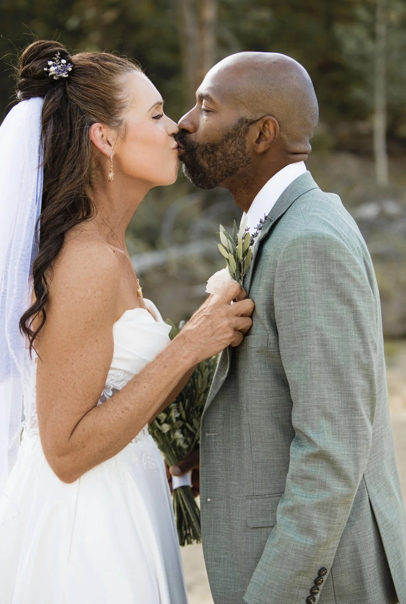 A bride and groom share a kiss outdoors, with trees in the background. The bride wears a white wedding dress and veil, and holds a bouquet. The groom wears a light gray suit.