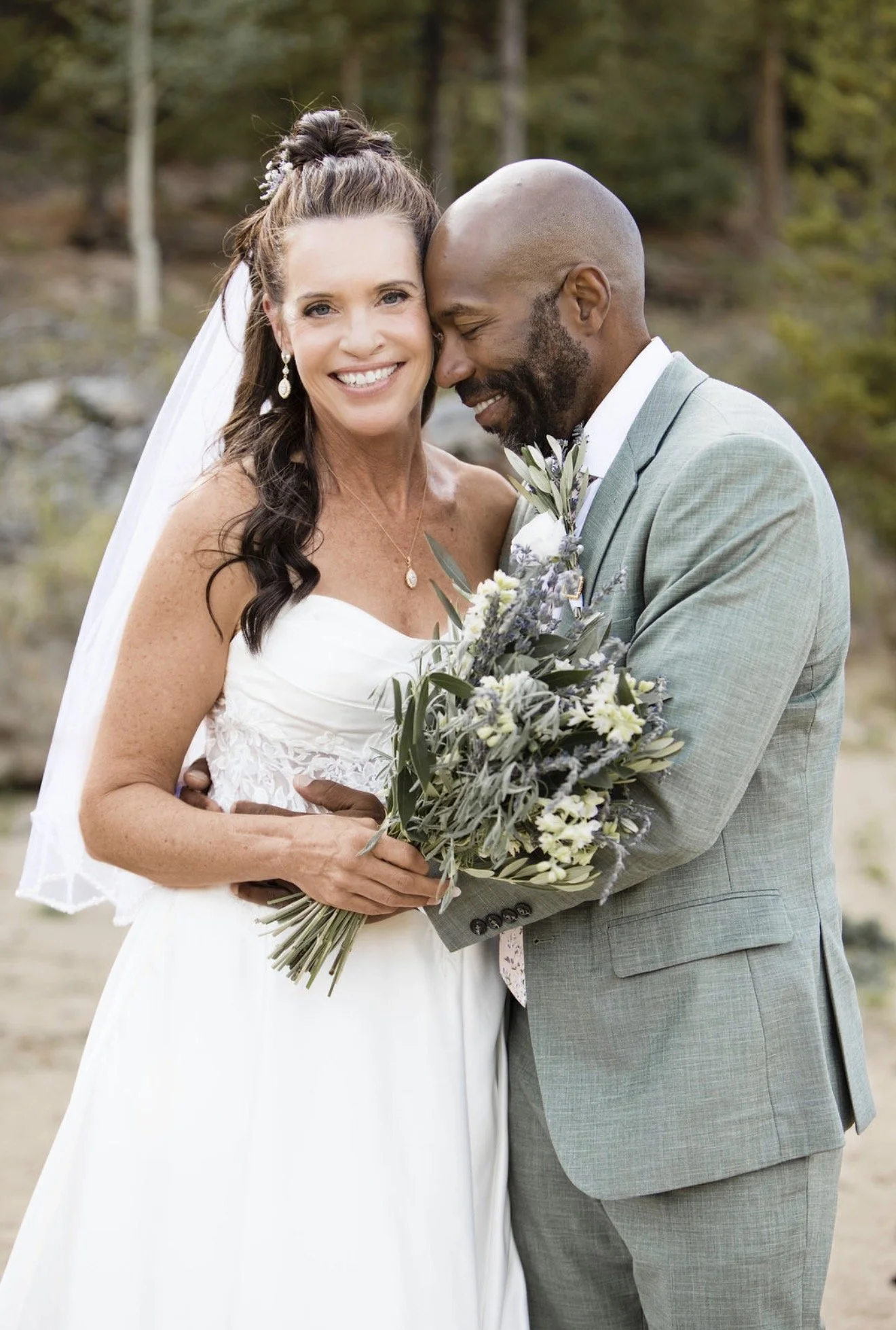 A bride and groom embrace outdoors, smiling, with the bride holding a bouquet of mixed flowers and greenery, both dressed in wedding attire.