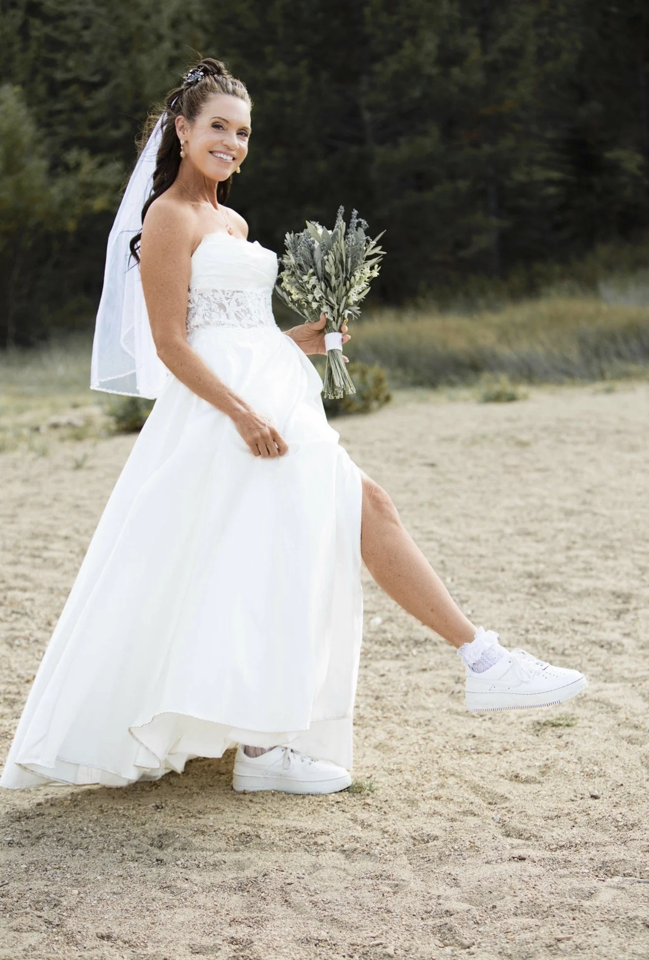A smiling woman in a wedding dress with a veil, holding a bouquet, and wearing white sneakers, outdoors on a dirt path with trees in the background.