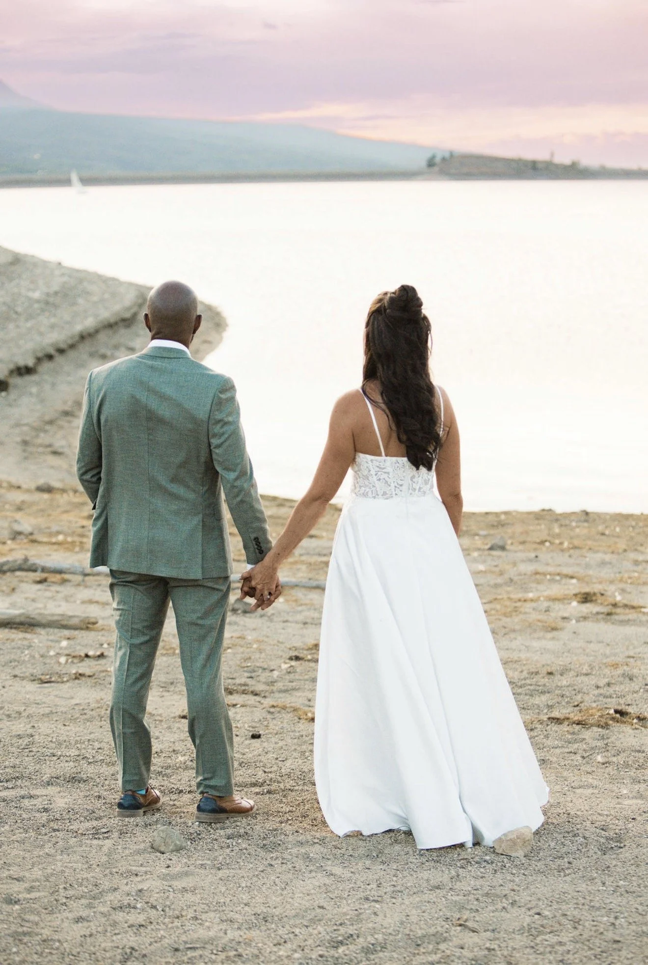 A couple dressed in wedding attire holding hands and walking along a rocky beach near a body of water during sunset.