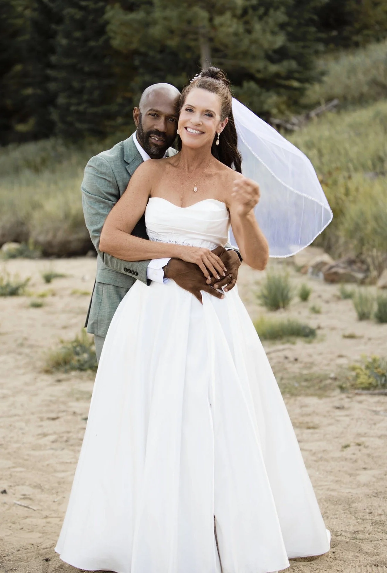 A happy bride and groom embracing outdoors, with a wedding dress, veil, and a suit in a natural setting with greenery.