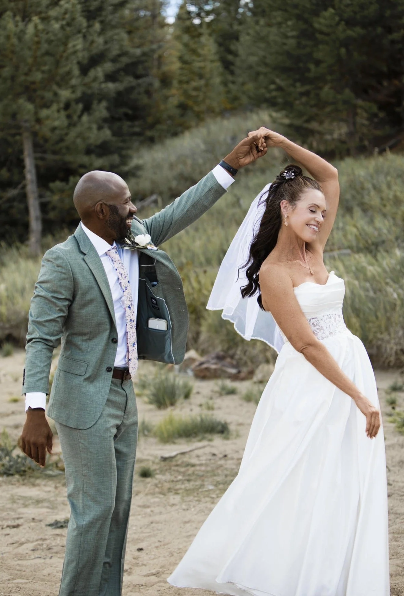 A man in a gray suit and a woman in a white wedding dress dancing outdoors in a sandy area with trees in the background.