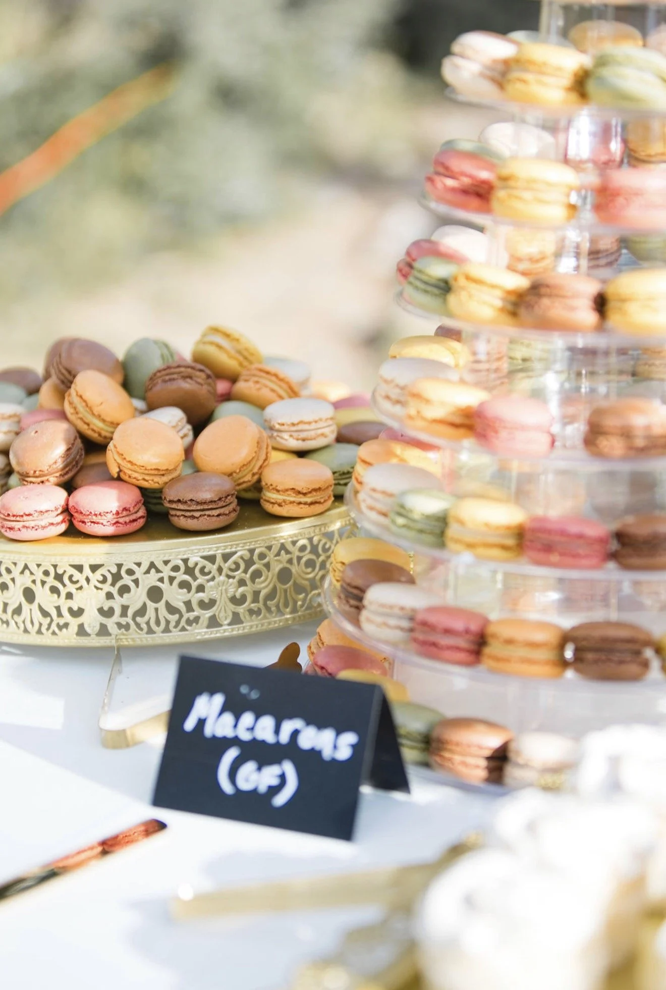 Colorful macarons on a white table with a gold decorative tray and a tiered stand, with a black chalkboard sign in the foreground that reads 'Macarons (GF)'.