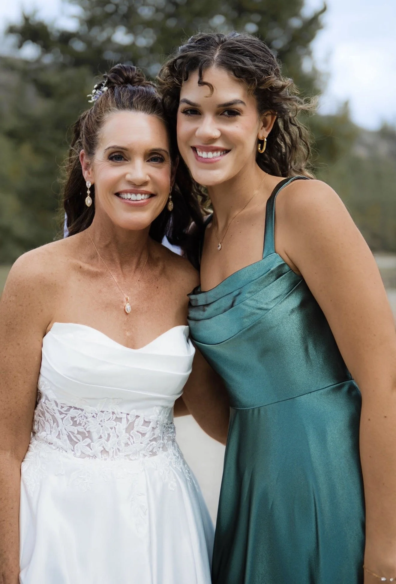 Two women are standing closely outdoors, smiling at the camera. One is wearing a white dress with lace detailing and jewelry, and the other is wearing a green satin dress with jewelry. They appear to be attending a formal event.