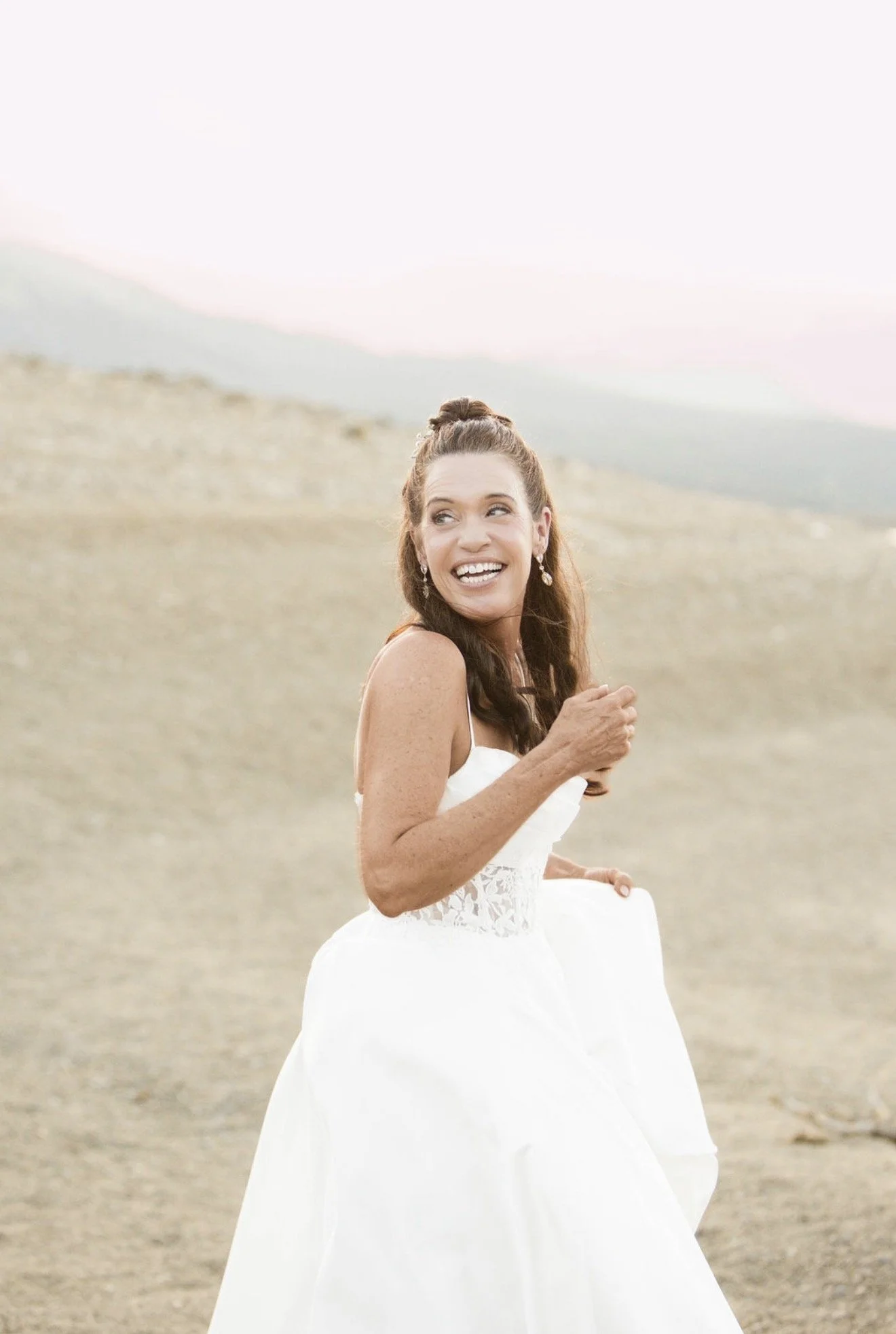 A woman in a white dress smiling and looking to the side in a desert landscape during sunset or sunrise.