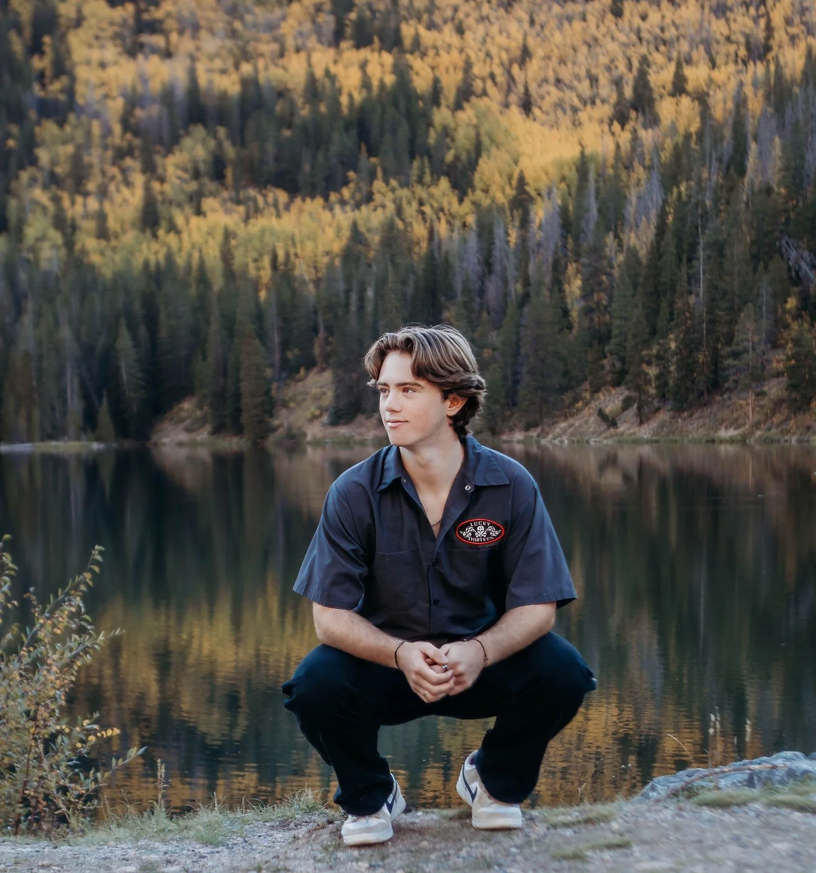 A young man with brown hair sitting outdoors near a lake with a forested mountain background, wearing a dark blue shirt and pants, and smiling while looking to the side.