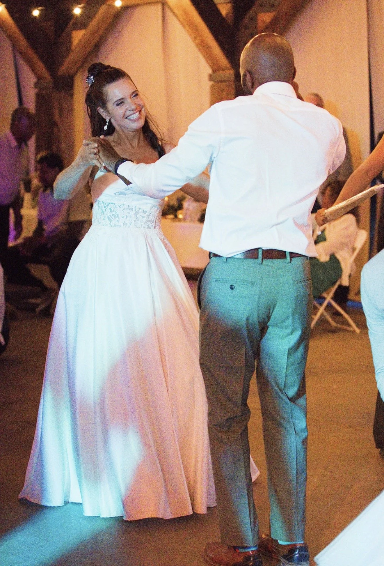 A woman in a white dress dancing with a man in a white shirt and gray pants at a wedding reception, indoors with wooden beams and other guests in the background.