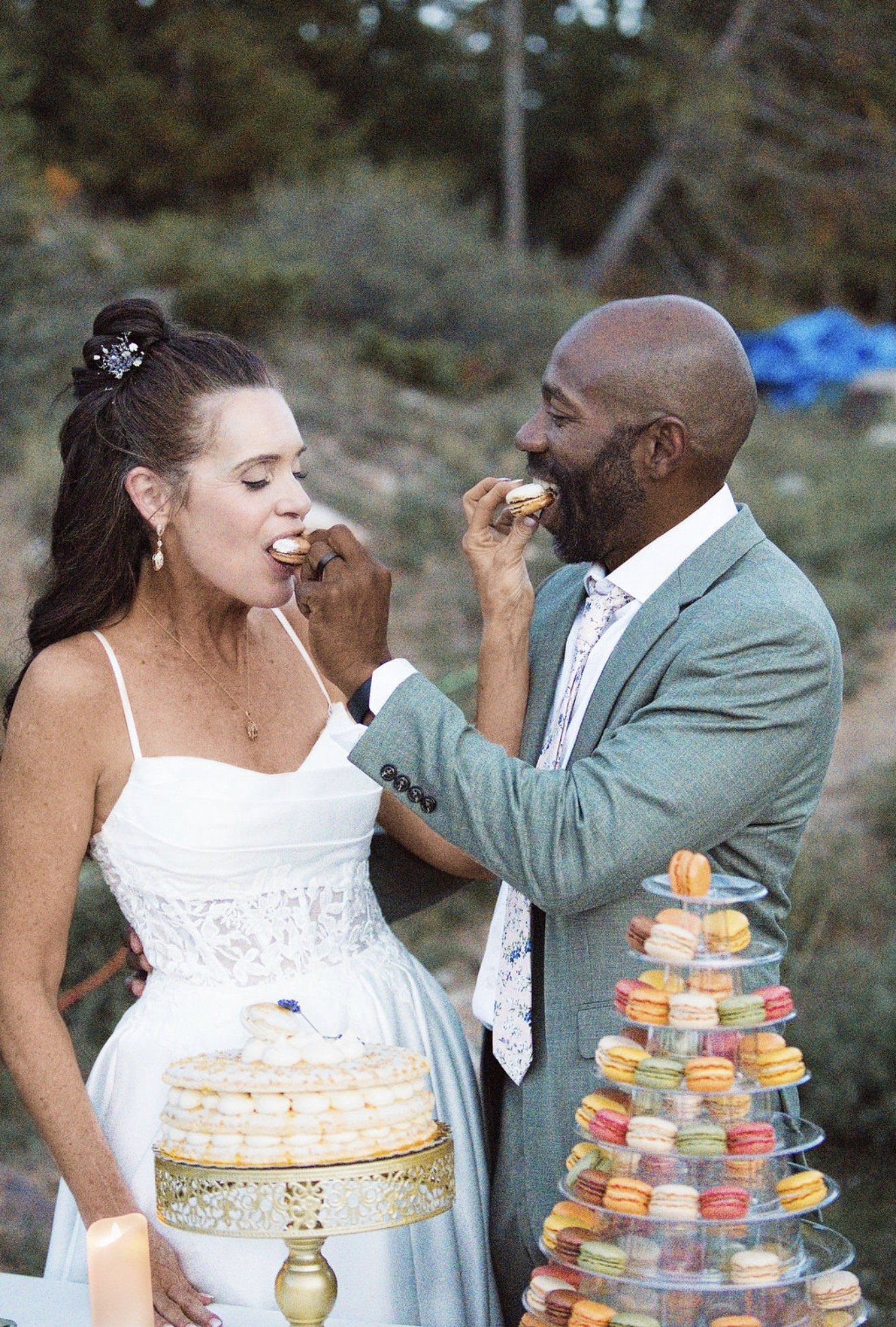 A bride and groom at a wedding cake and macarons dessert table, feeding each other macarons outdoors amid greenery.