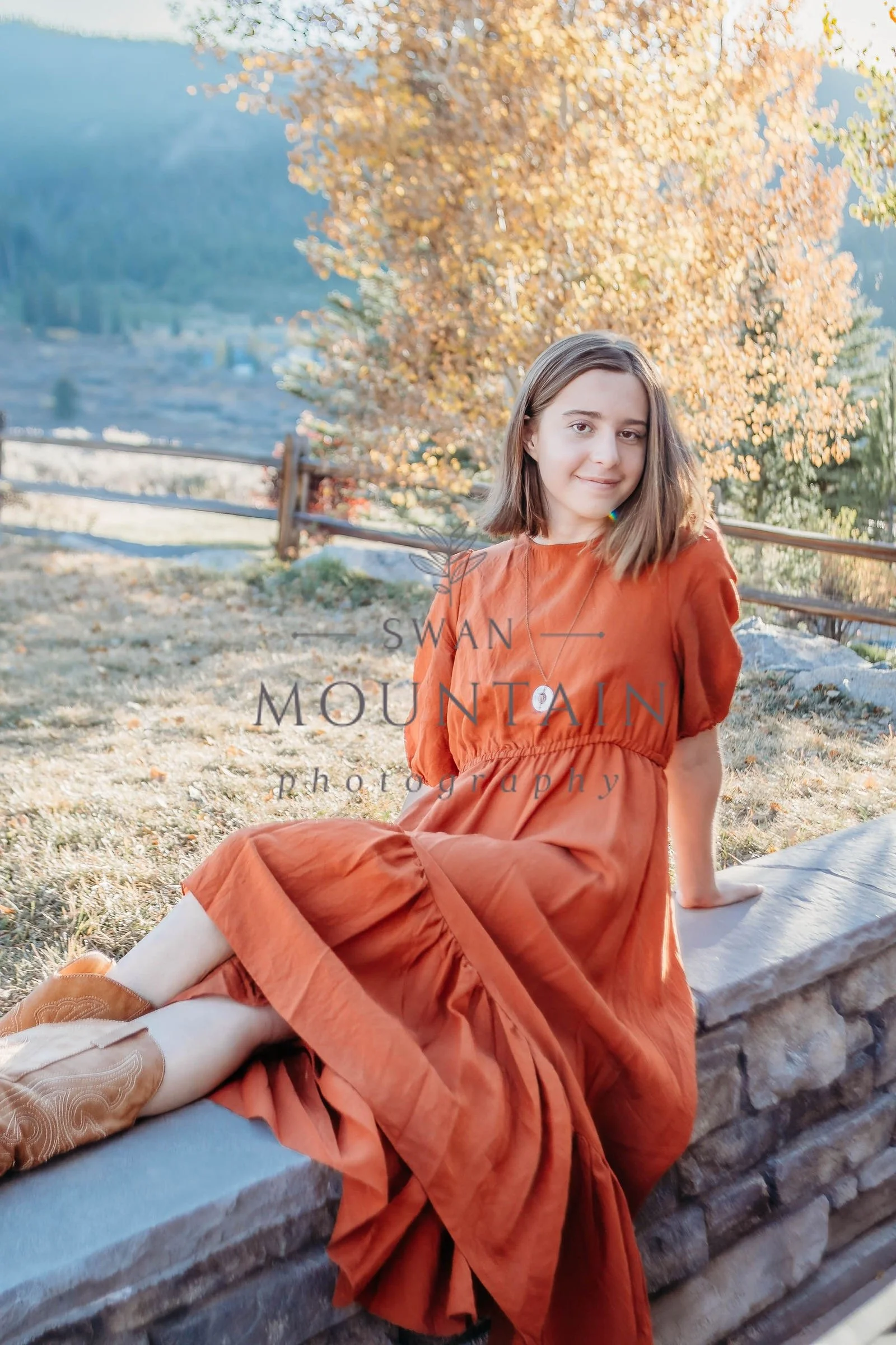 A young woman with shoulder-length brown hair sitting on a log outdoors near a tree with orange and yellow leaves, on a sunny day in autumn.