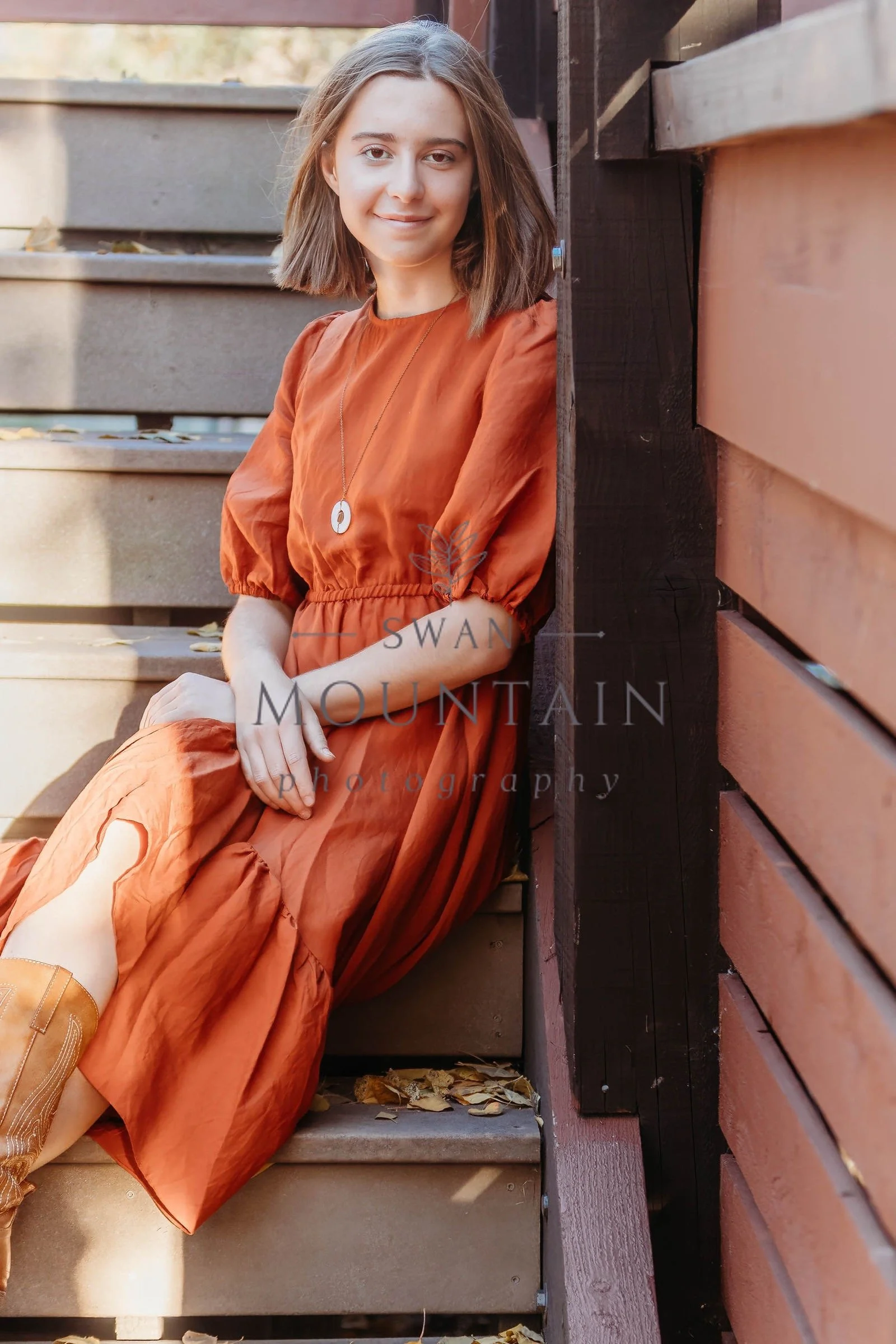 Young woman with shoulder-length brown hair, wearing a rust-colored dress and a necklace, sitting on stairs outside next to a wooden railing, with fallen leaves around.