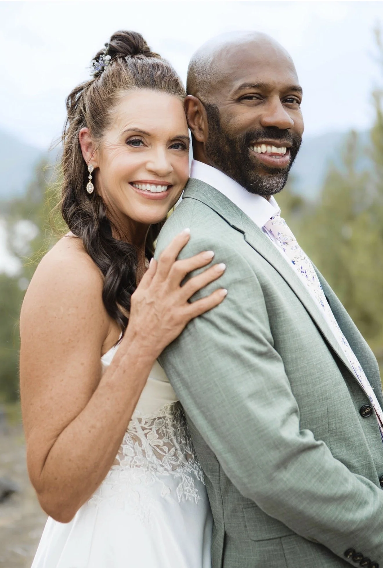 A smiling couple, a woman with long dark hair styled in a bun with floral accessories, wearing pearl earrings and a white wedding dress, and a man with a beard in a light gray suit, standing outdoors with a blurred natural background.
