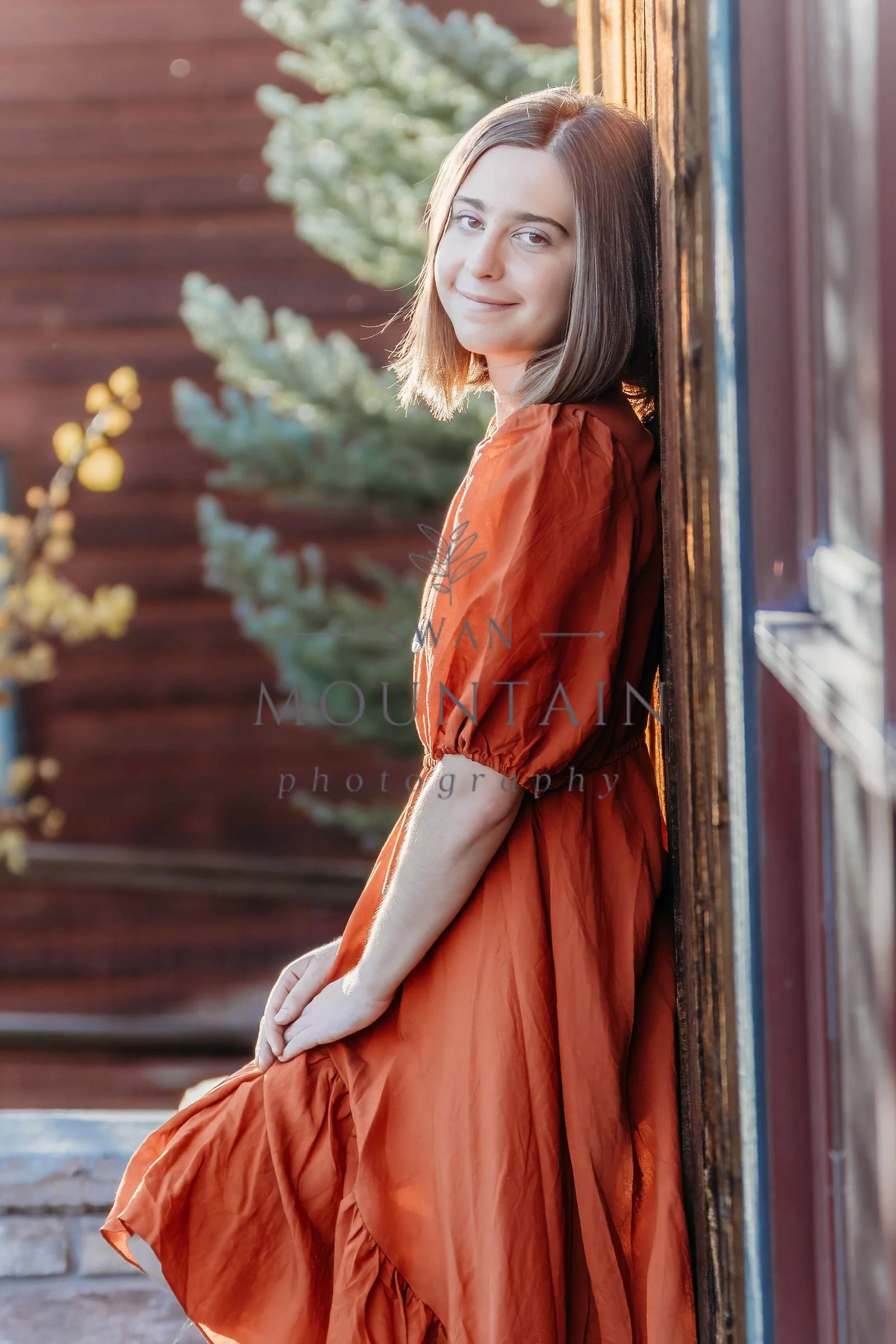 A young woman with shoulder-length brown hair leaning against a wooden wall outside, wearing an orange dress with puffed sleeves. She is smiling softly, with sunlight illuminating her face. In the background, blurred greenery and wooden stairs are vi