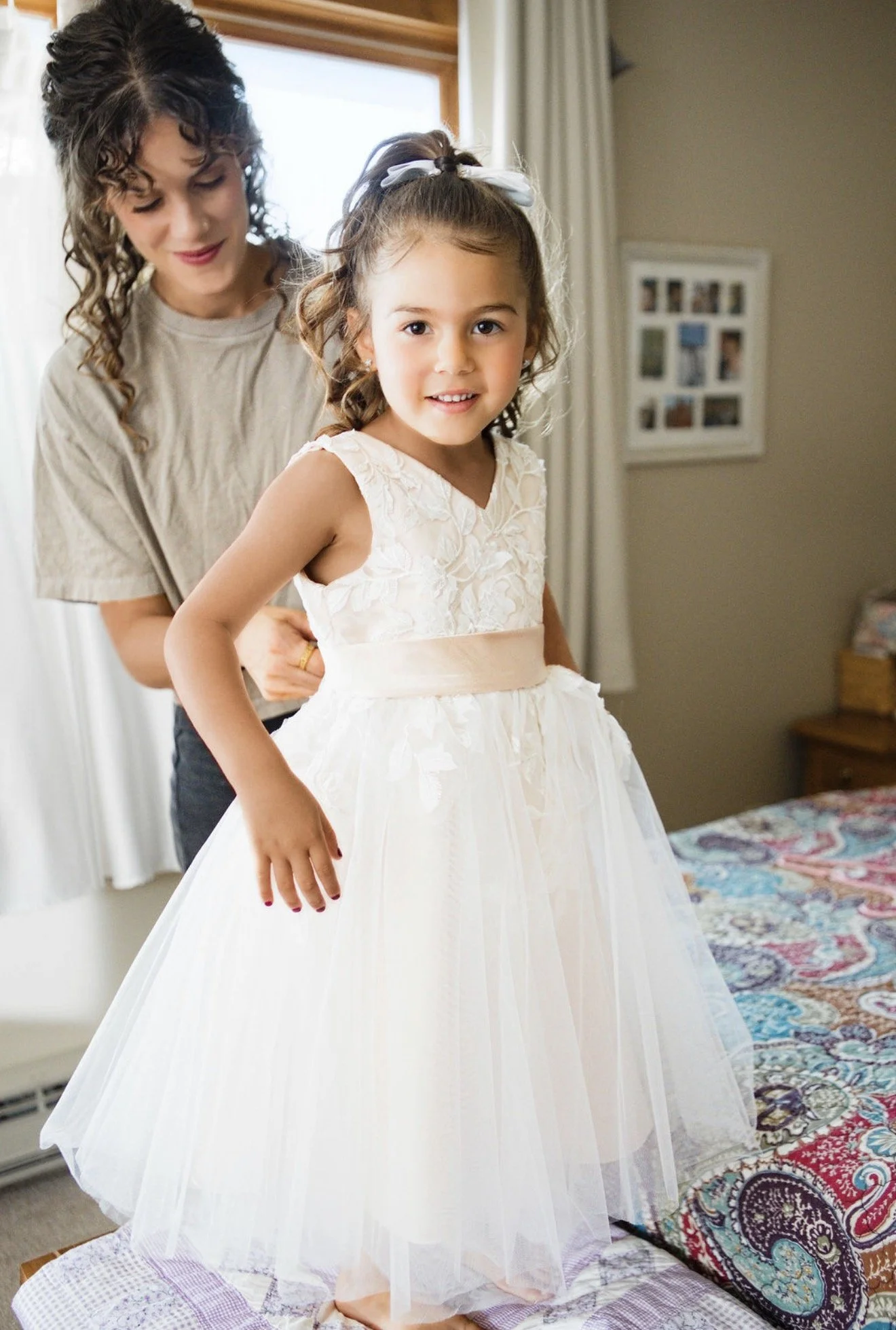 A young girl wearing a white princess dress with floral details and a white bow in her hair, standing on a bed with her mother helping her get ready, in a bedroom with a window and framed photographs on the wall.