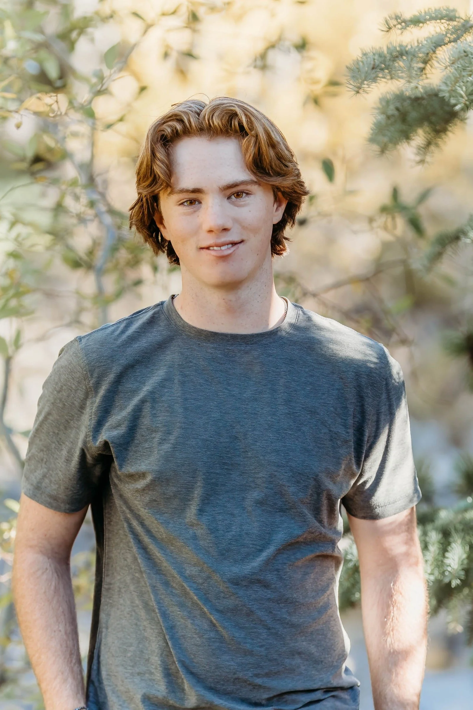 A young man with reddish hair and freckles wearing a gray T-shirt standing outdoors among green foliage with sunlight filtering through the leaves.