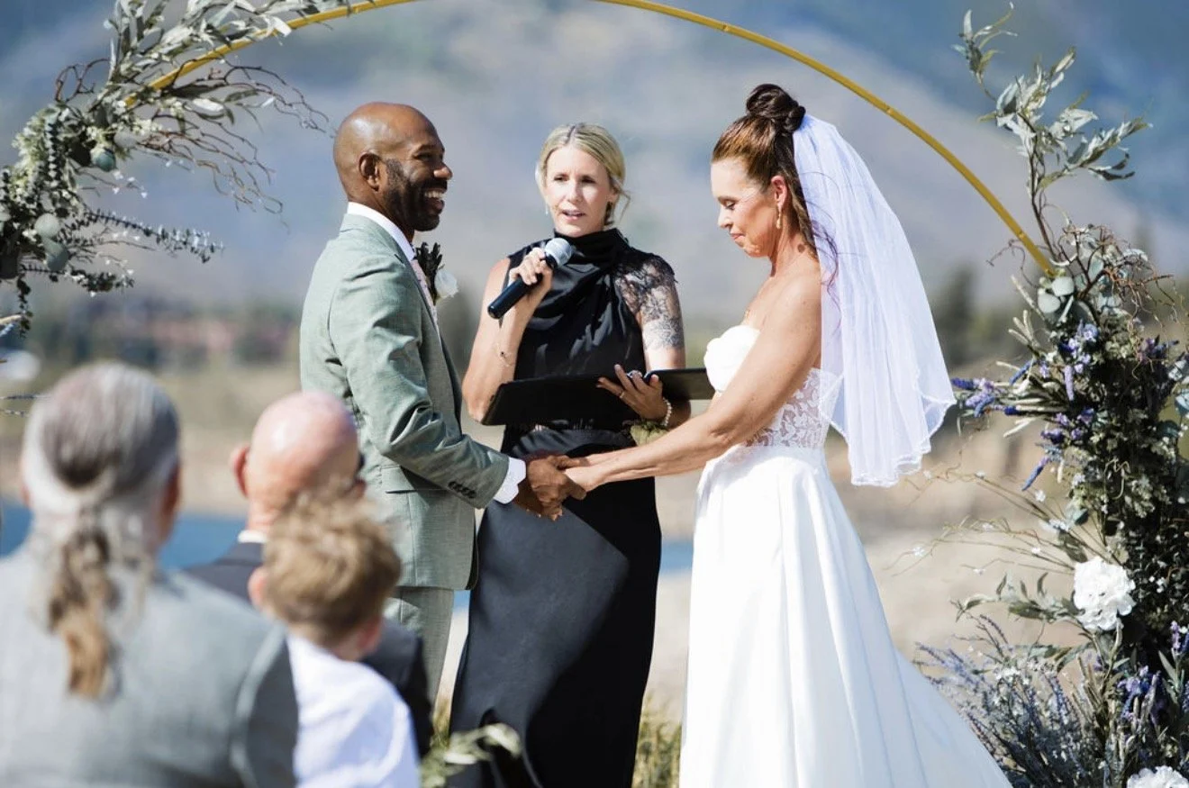 A couple getting married outdoors, holding hands and exchanging vows, with an officiant reading as guests watch, floral arch backdrop, mountains in the background.