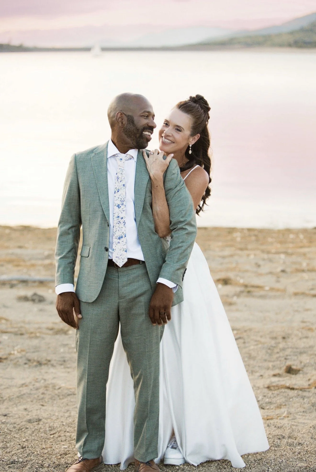 A couple in wedding attire standing on a beach at sunset, smiling and looking at each other, with sailboats and water in the background.
