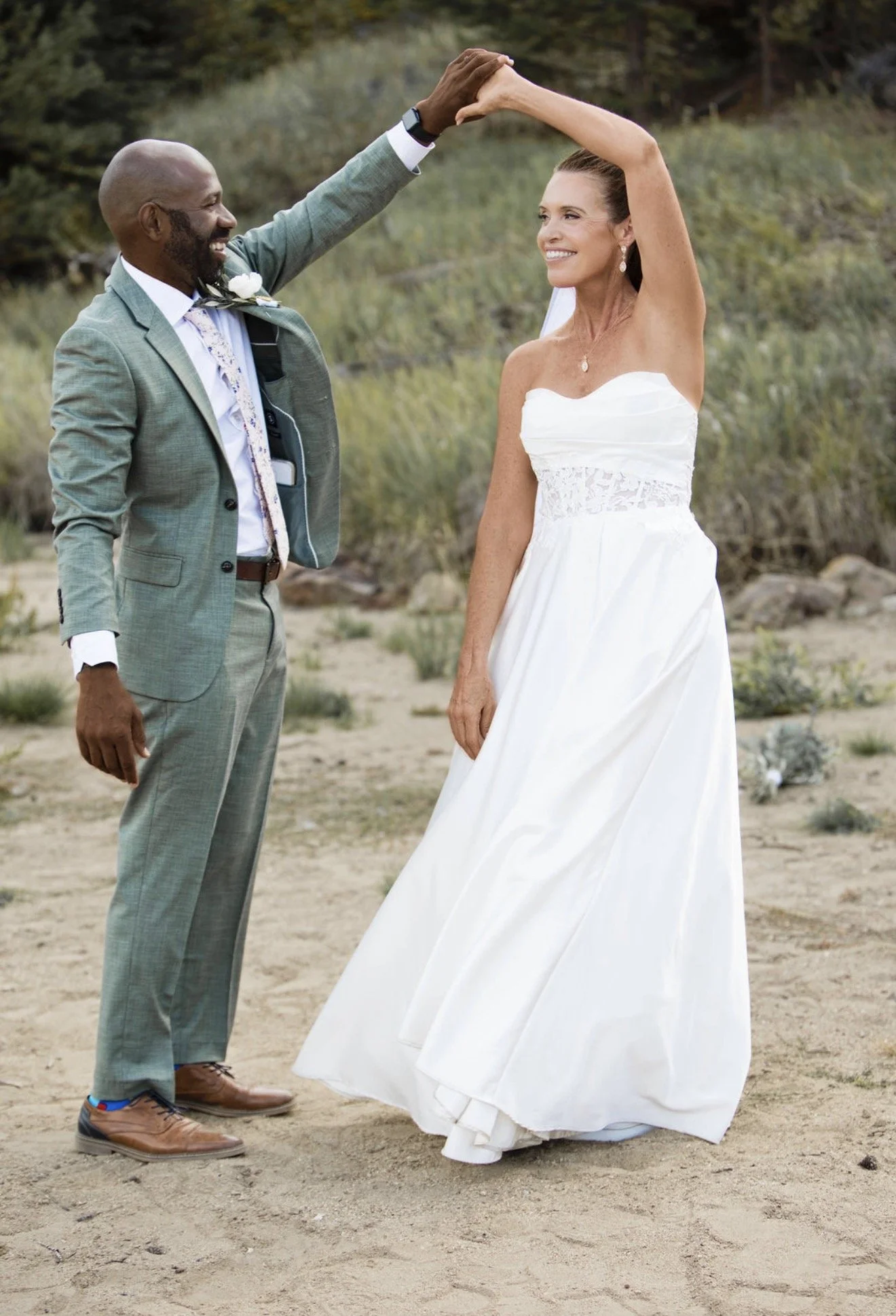 A couple, the man with dark skin and the woman with light skin, dancing outdoors in a natural setting, the man wearing a green suit and the woman in a white wedding dress, smiling at each other.