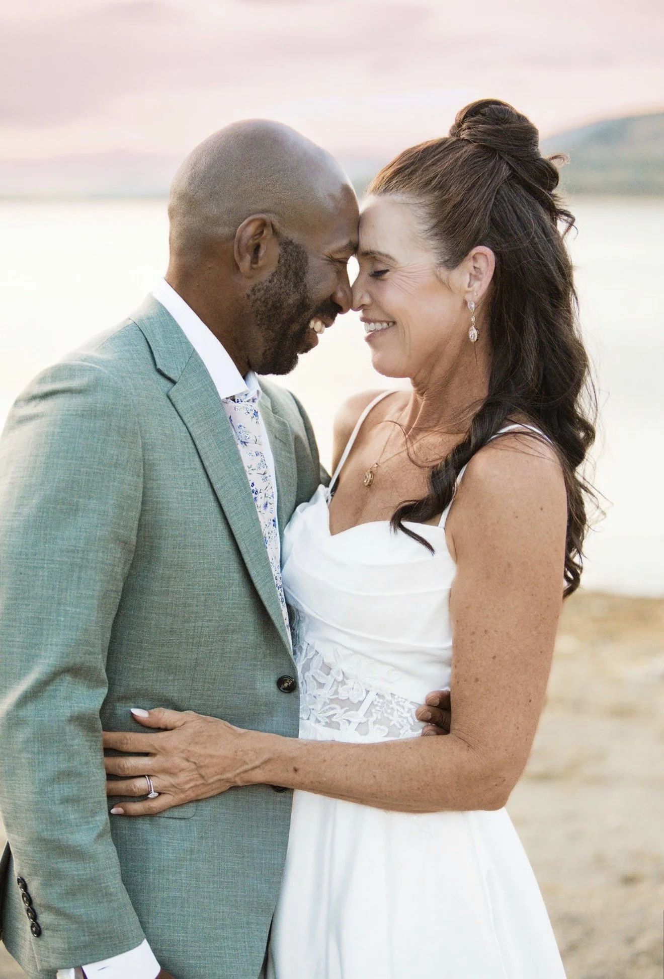 A couple in wedding attire touching foreheads and smiling on a beach at sunset.