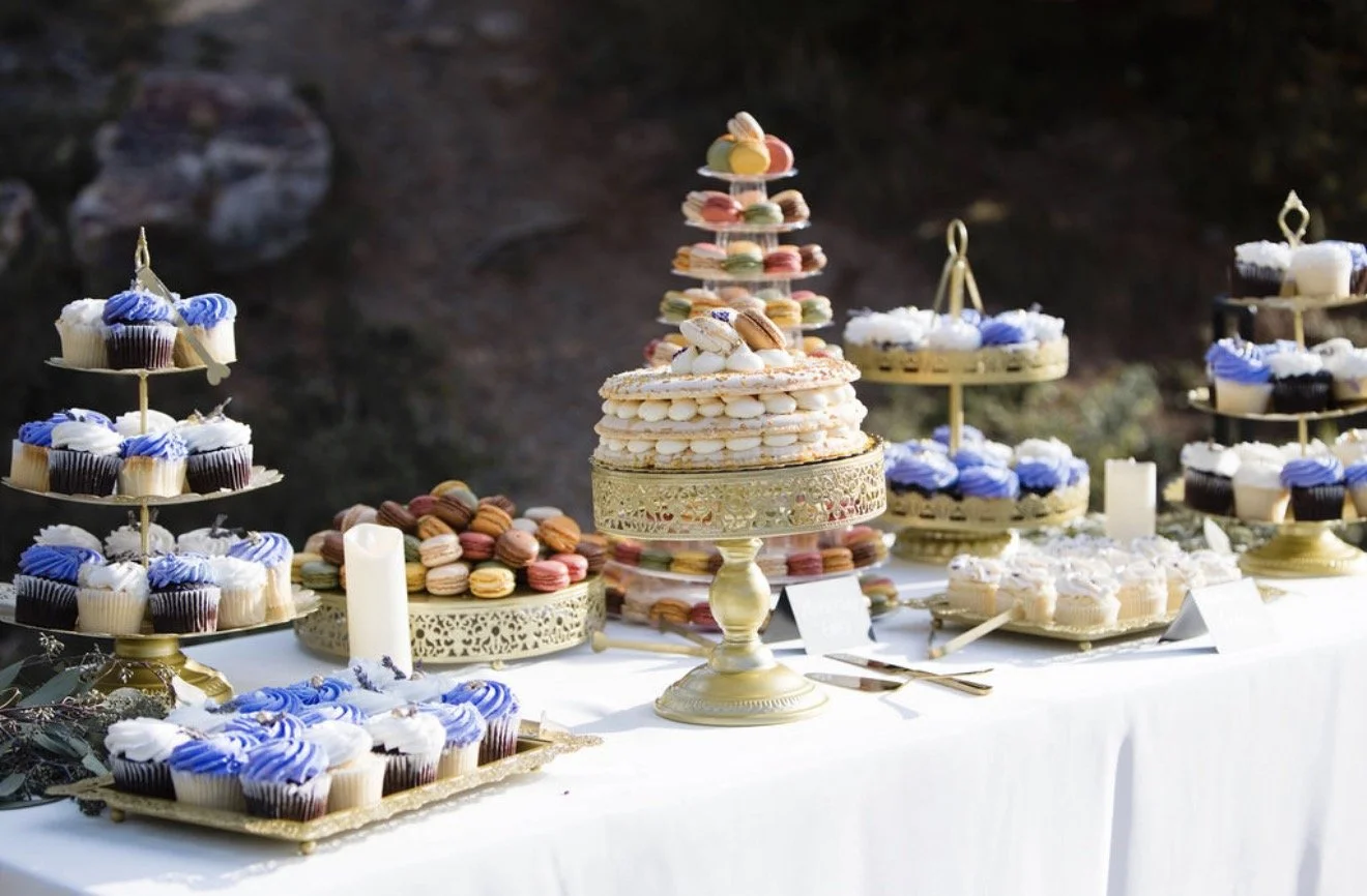 Table with an assortment of cupcakes, macarons, and a layered cake for a celebration, with decorative candles and greenery.