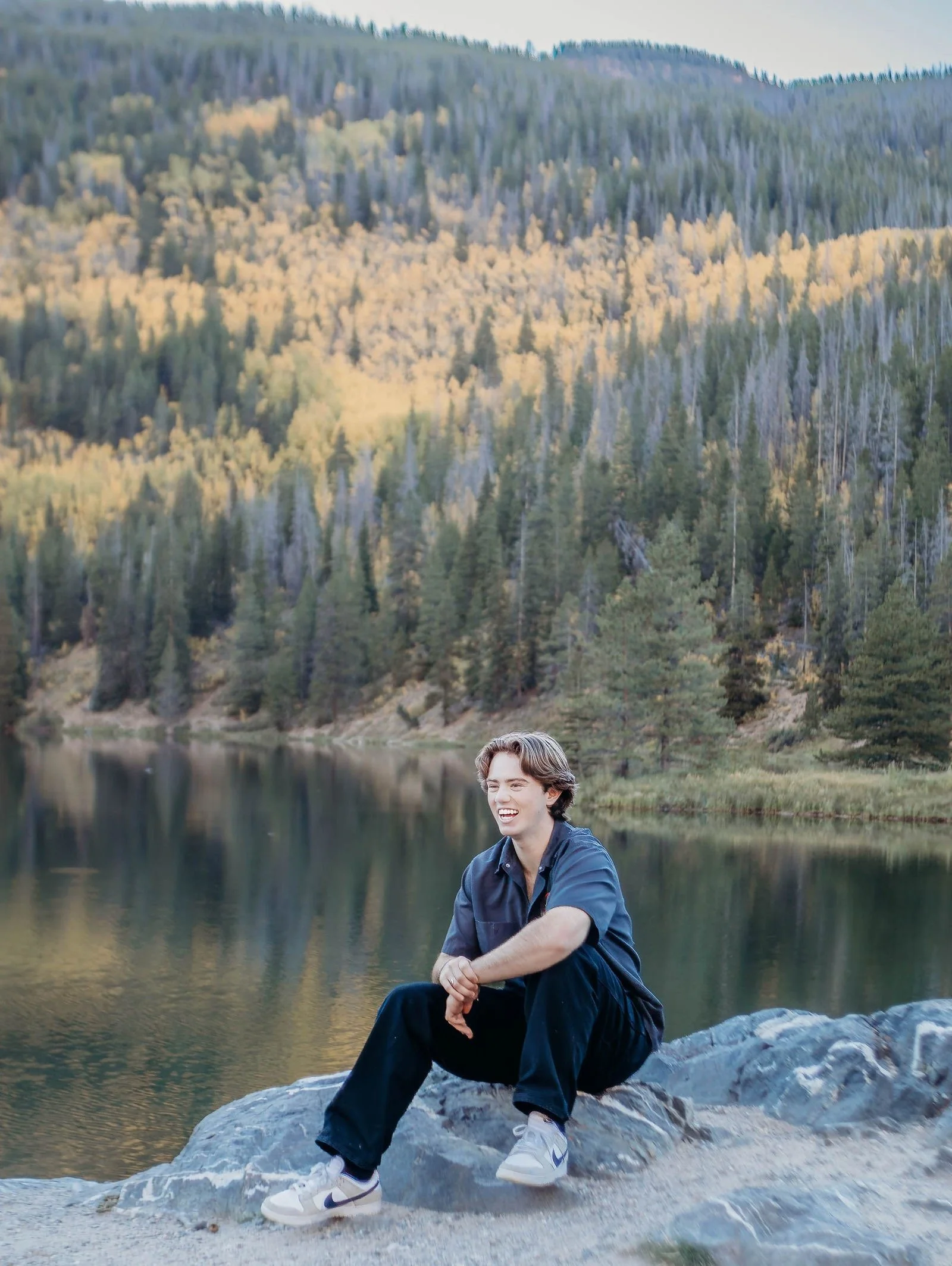 A young person with brown hair sitting on a rock by a lake, smiling, with a forested mountain landscape in the background.