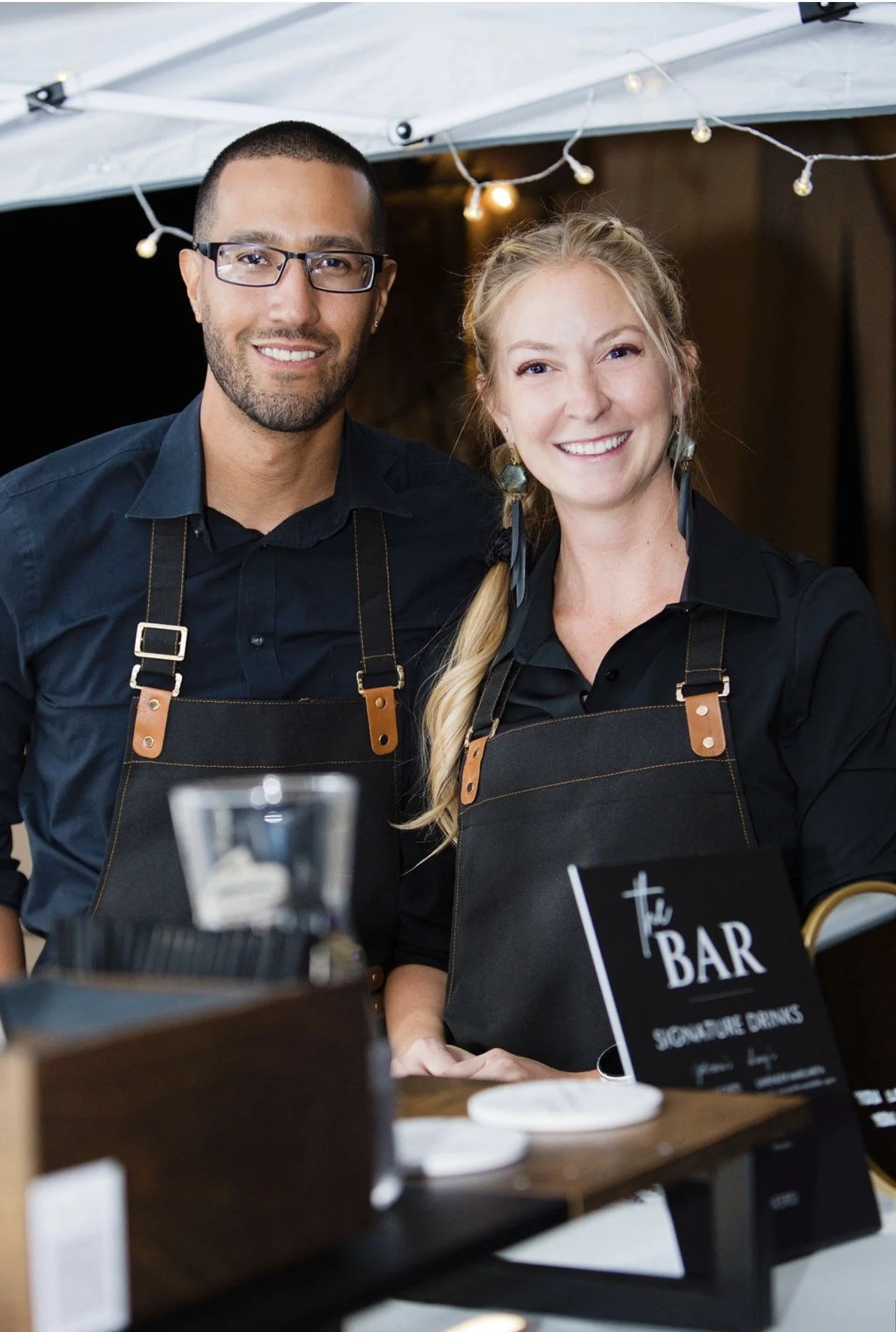 Two smiling people, a man with glasses and a woman with braided hair, standing next to each other behind a table at a bar with a sign that says "The Bar". They are wearing black shirts and aprons, with string lights hanging above them.