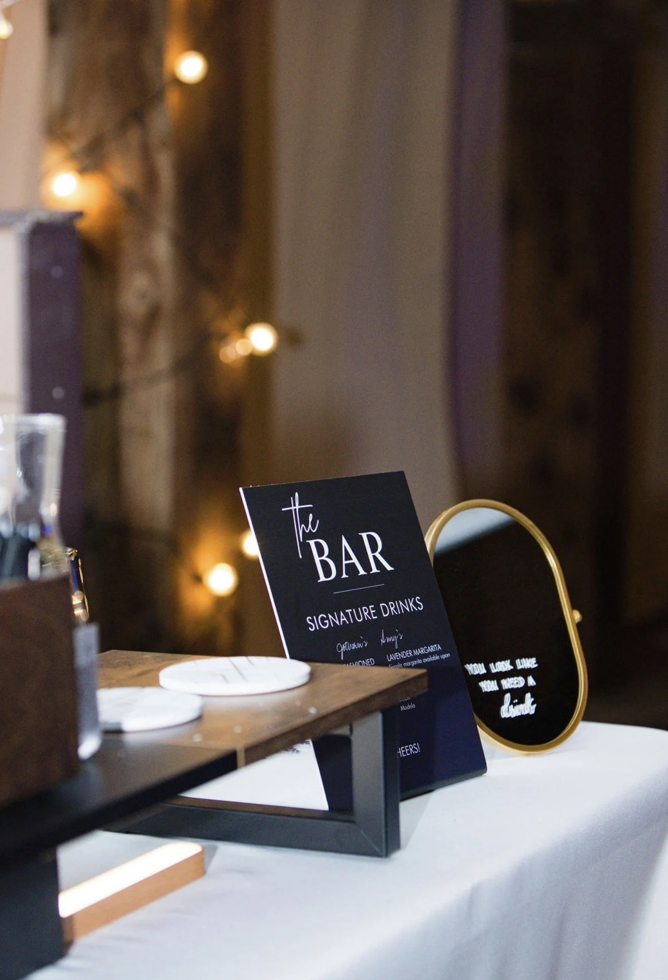 Close-up of a sign on a table that reads 'the BAR, signature drinks,' with a black background and white and purple text, at an event or wedding reception with blurred background lighting