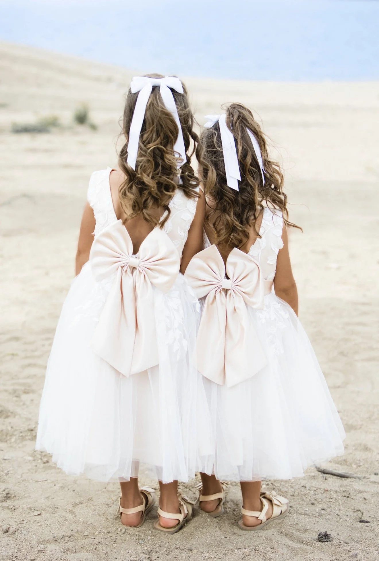 Three young girls dressed in white dresses with large bows on their backs, walking on a sandy beach, holding hands, with the ocean and sky in the background.