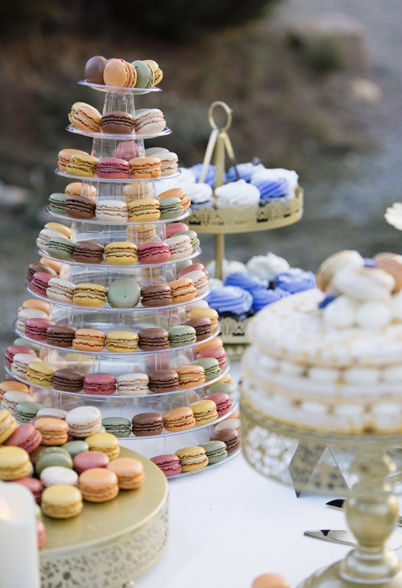 Display of colorful macarons arranged on multiple tiers of clear stands at a dessert table outside.