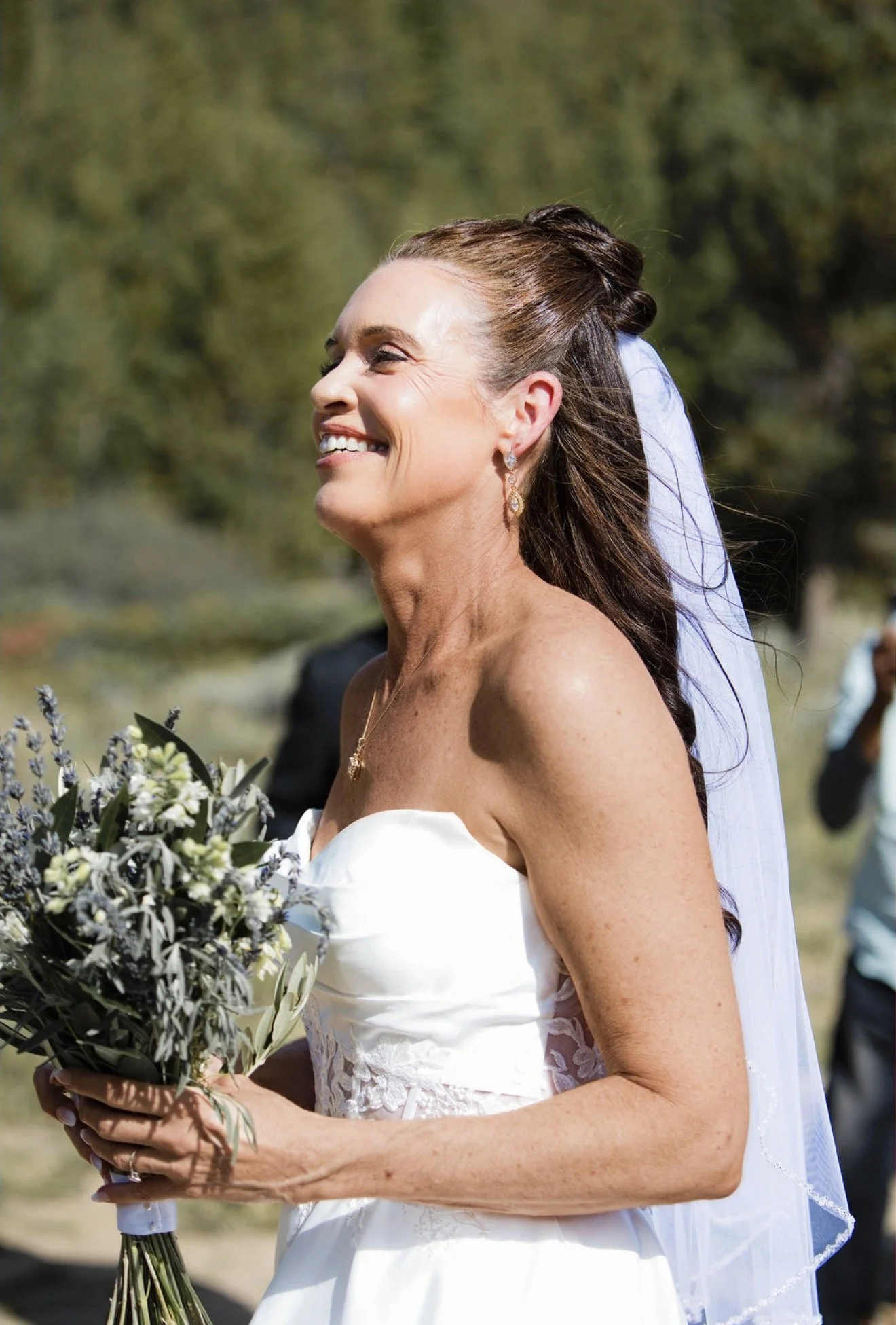 A smiling woman in a white wedding dress holding a bouquet of flowers at an outdoor wedding ceremony.